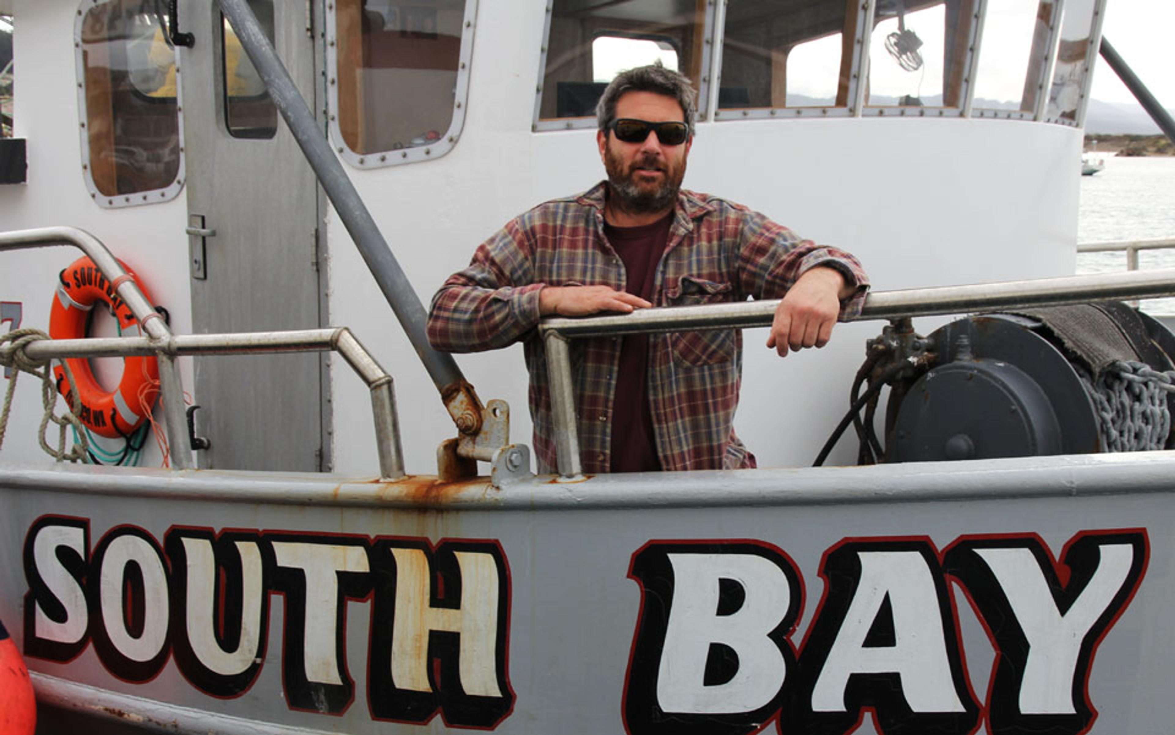 A man in a flannel shirt and sunglasses stands on a boat named “SOUTH BAY” with a visible lifebuoy.