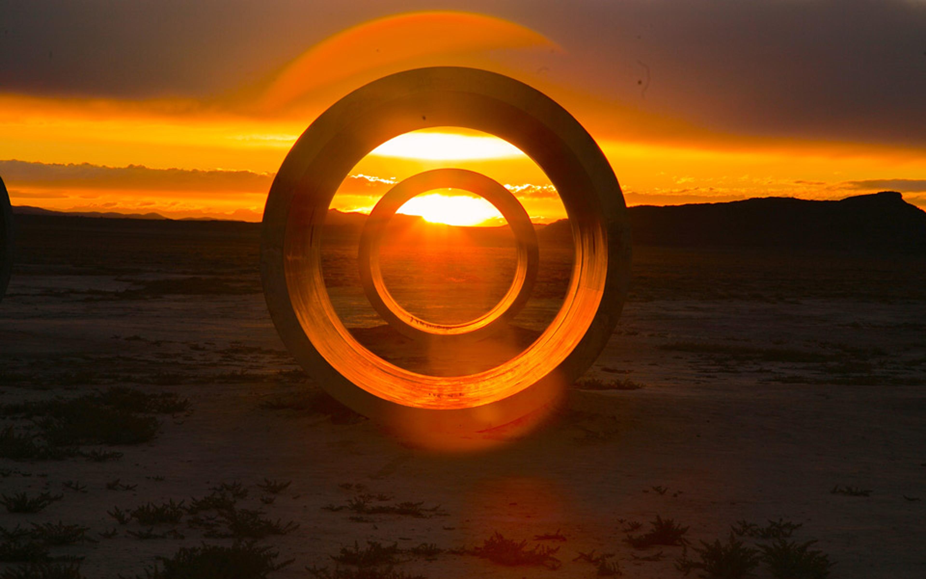 Photo of sunrise seen through large circular sculptures in a desert landscape, with an orange sky and distant mountains.