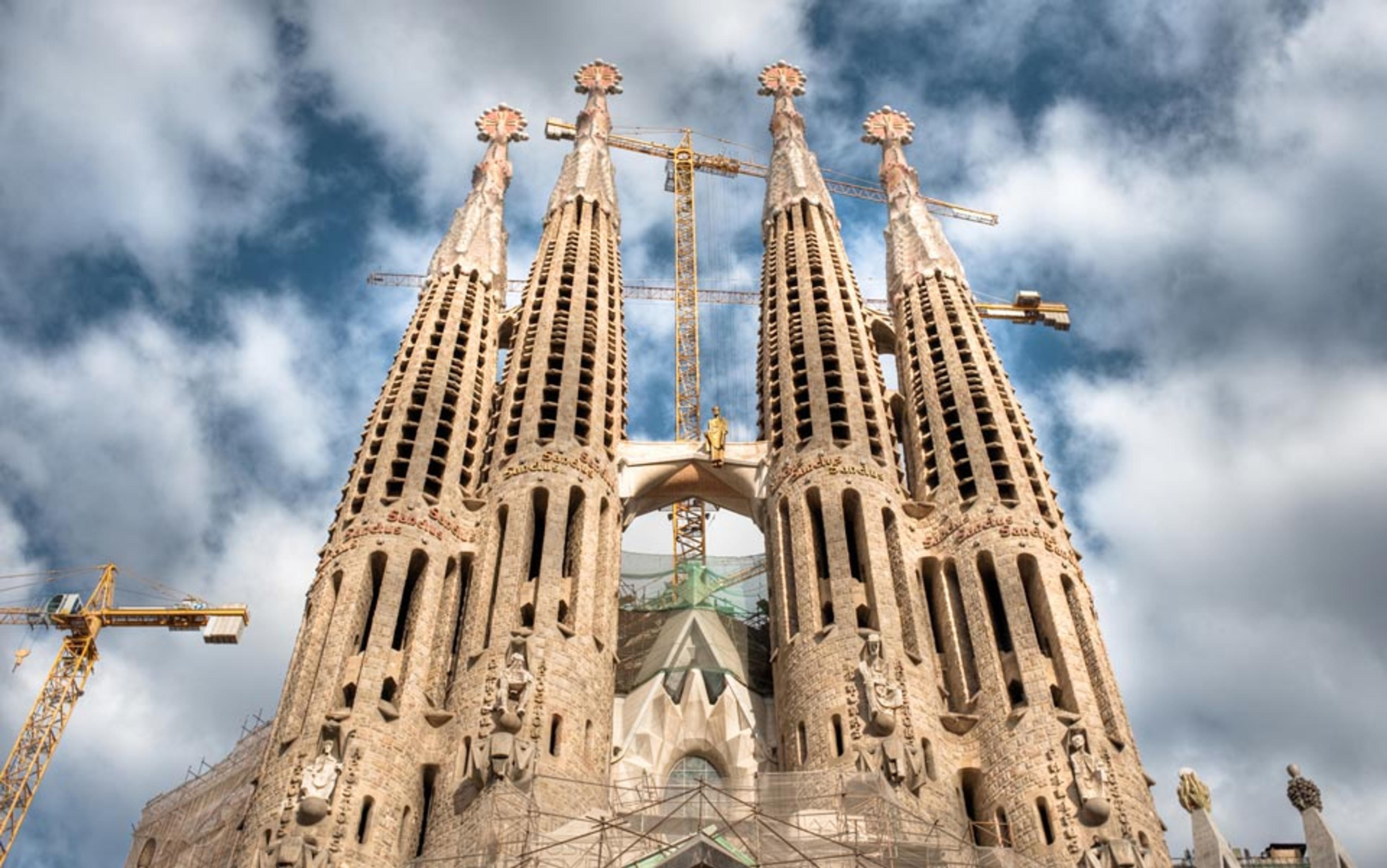 Sagrada Familia in Barcelona with four spires in the foreground, cranes around it, and a partly cloudy sky in the background.