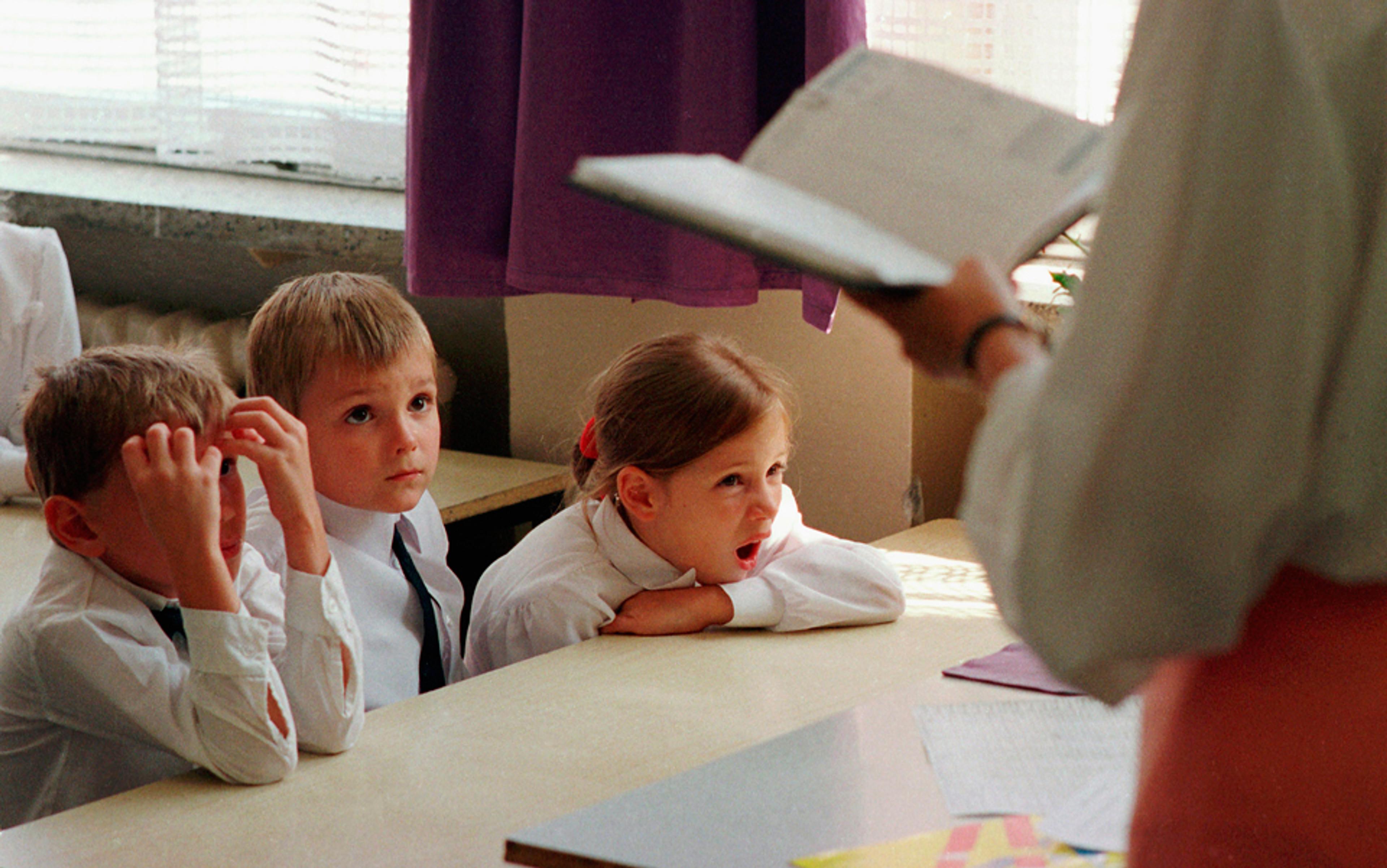 Photo of three young school children in uniform sitting at a desk with a teacher holding a book, one child yawning.