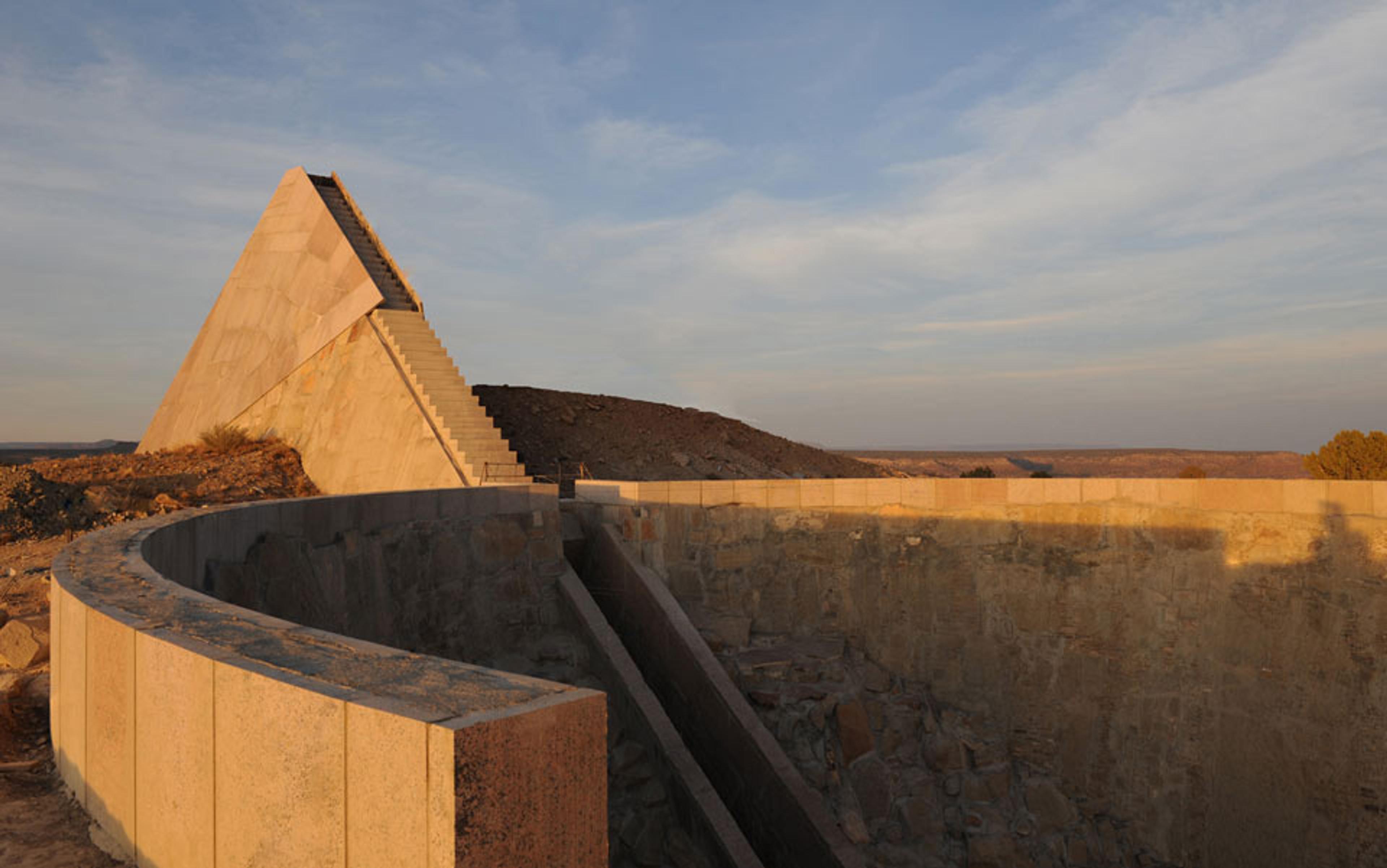 Photo of a modern stone pyramid with stairs, set in a desert landscape, with curved stone walls in the foreground and a cloudy sky.