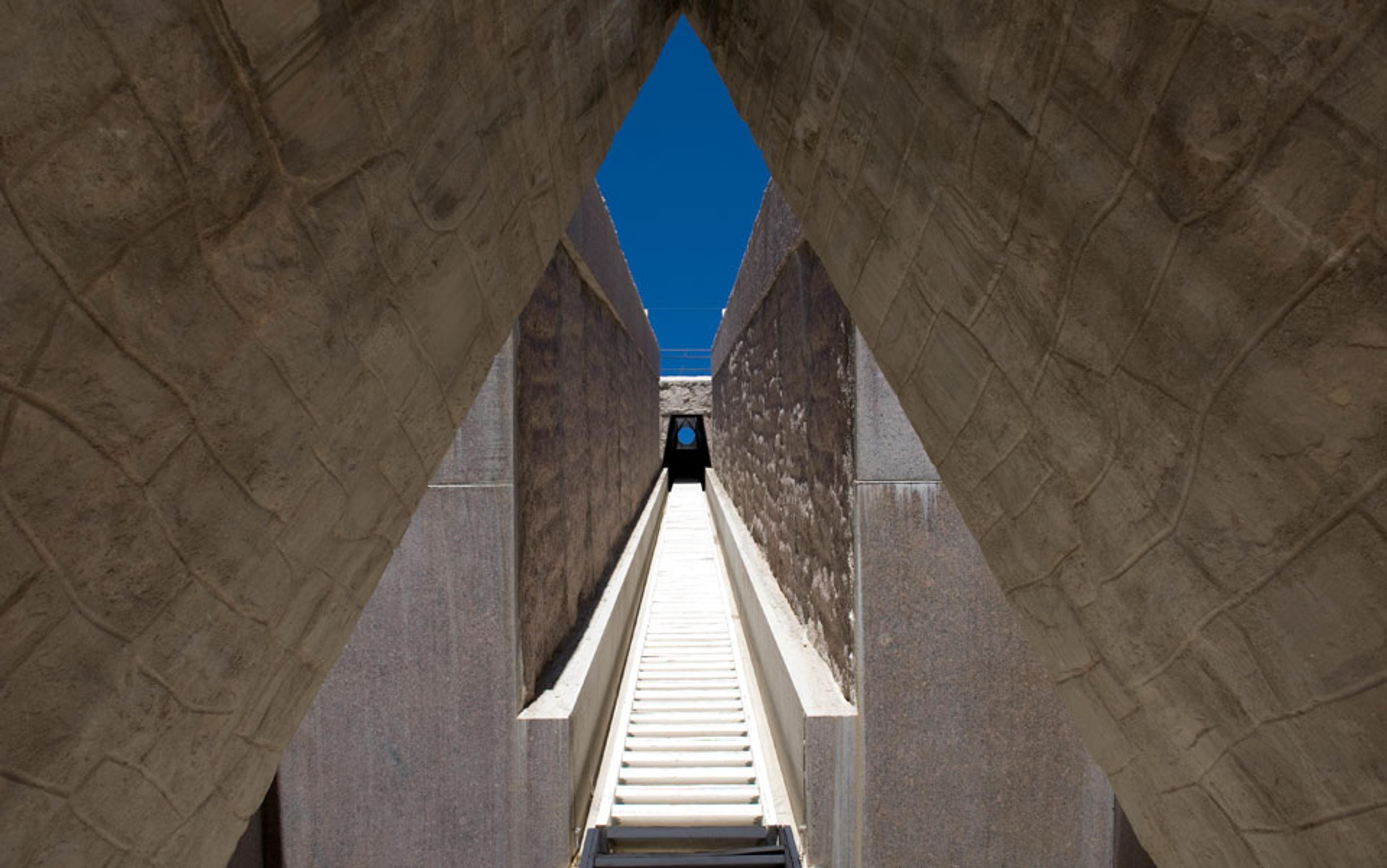 Photo of a narrow stone pathway between high walls leading to a small, distant archway under a clear blue sky.