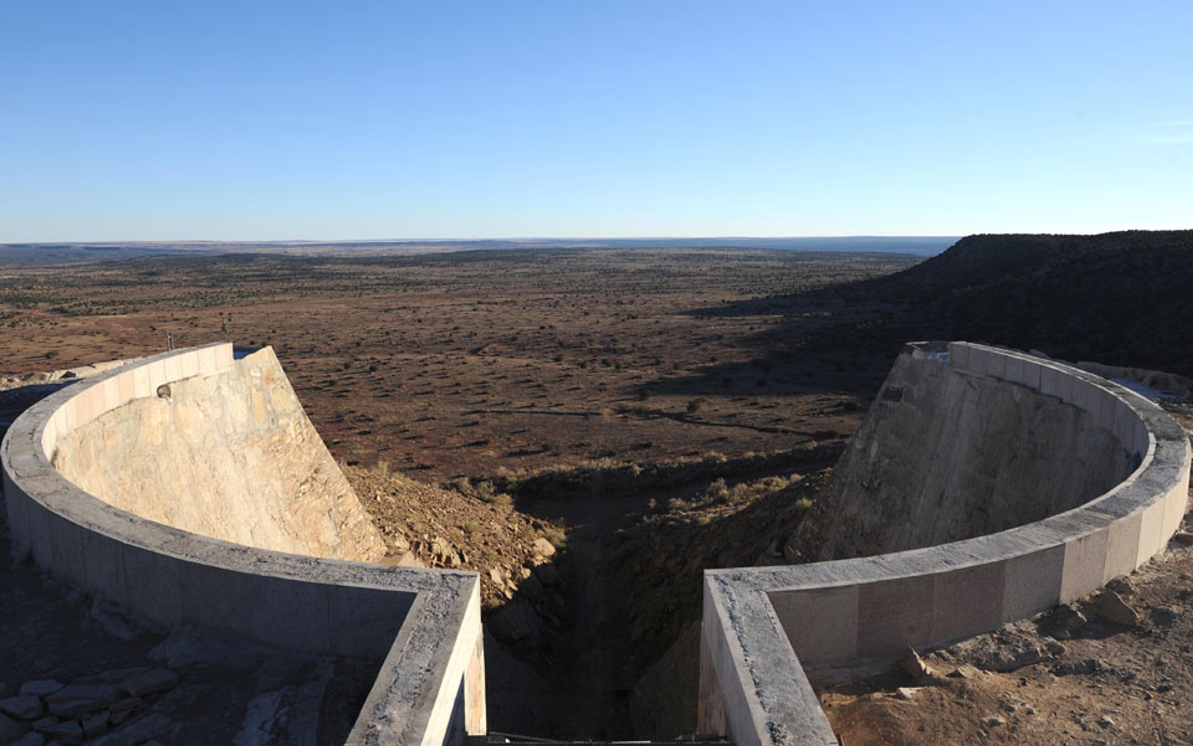 Photo of a dry landscape seen from behind a semi-circular dam structure, with a clear blue sky overhead.