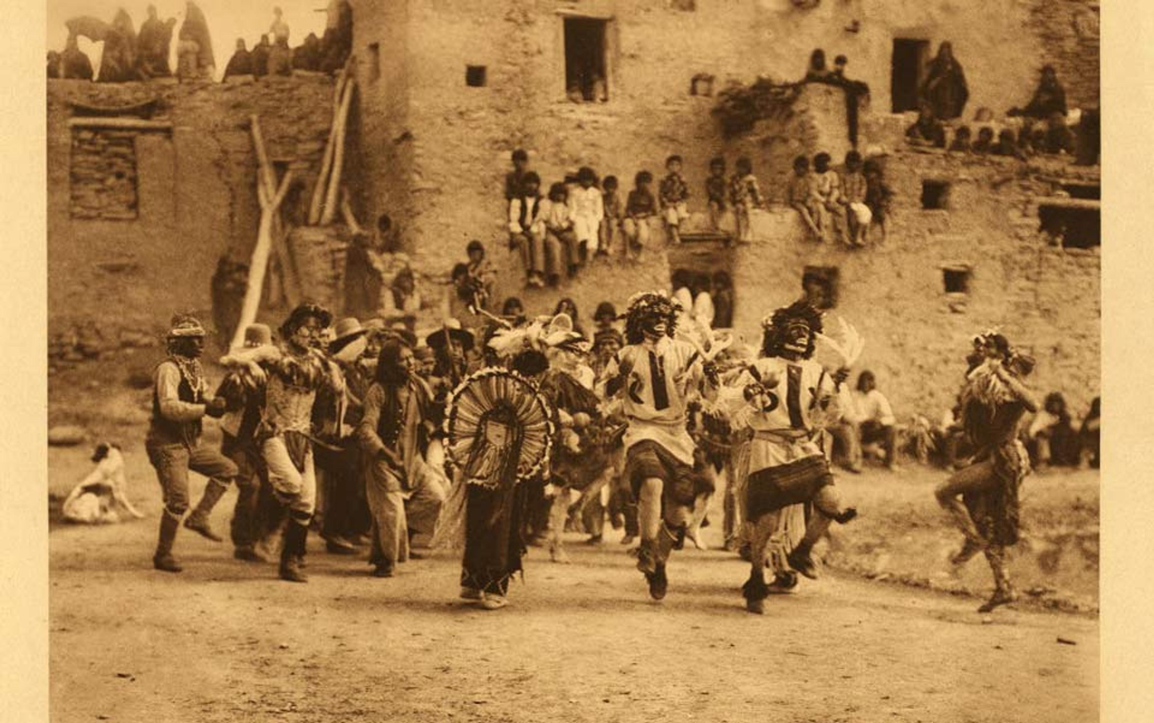 Sepia-toned photo of a traditional dance performed by people in masks and costumes in front of an adobe dwelling with onlookers.