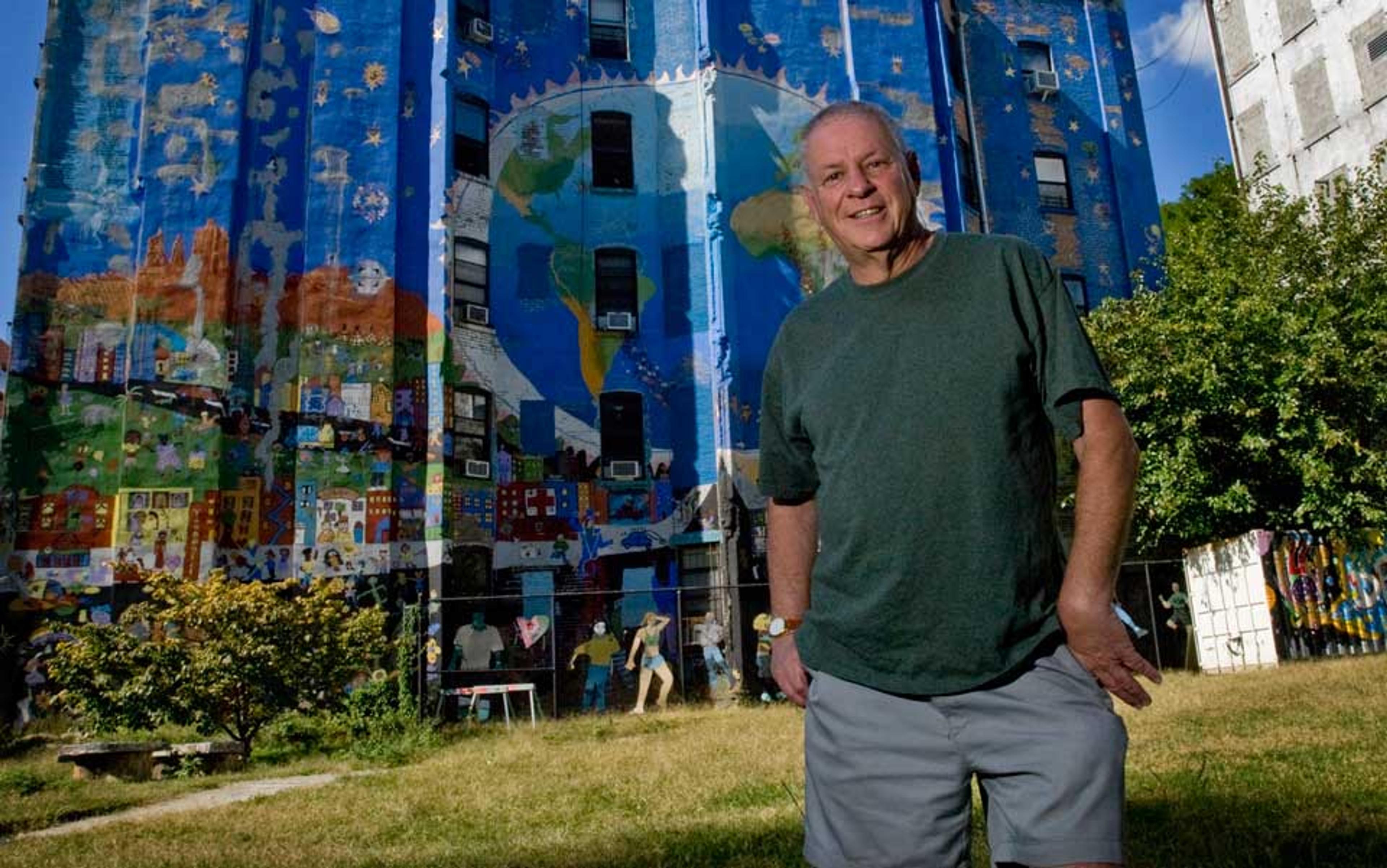 A man in a green shirt and shorts standing in front of a brightly painted mural on a tall building depicting a cityscape and Earth.