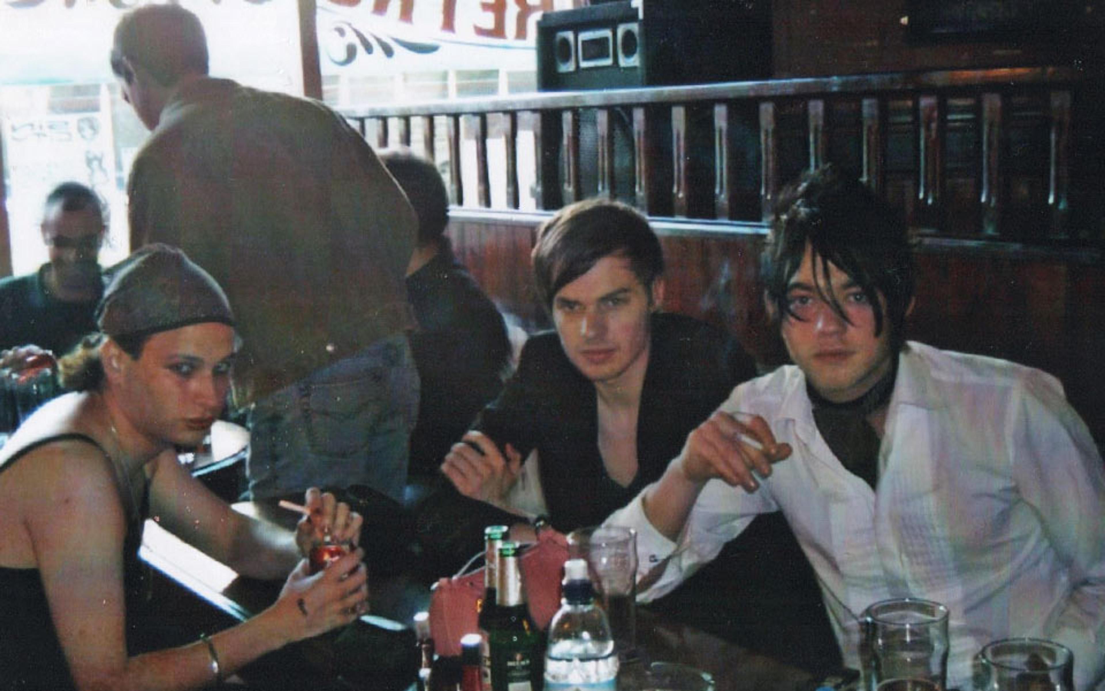 Three people sitting in a dimly lit pub with drinks on the table. A fourth person stands in the background.