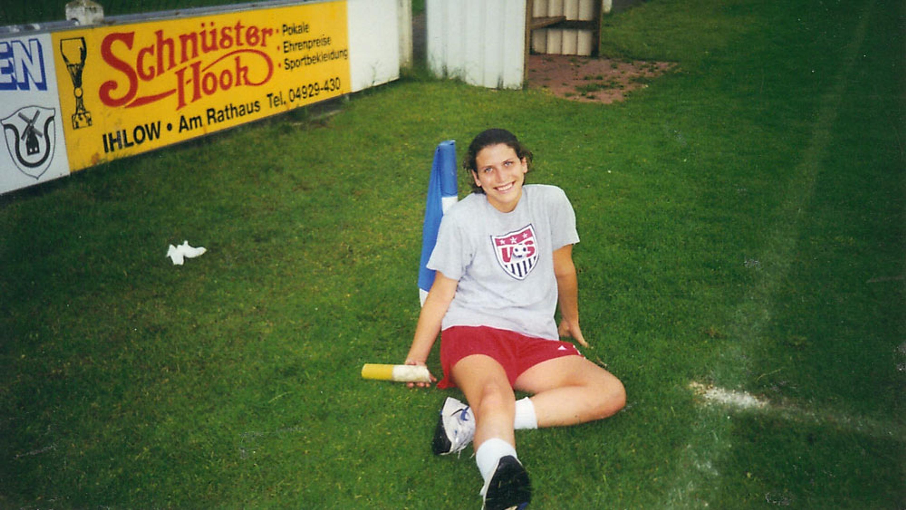 A smiling woman sitting on grass at a sports field wearing a US soccer shirt and red shorts.