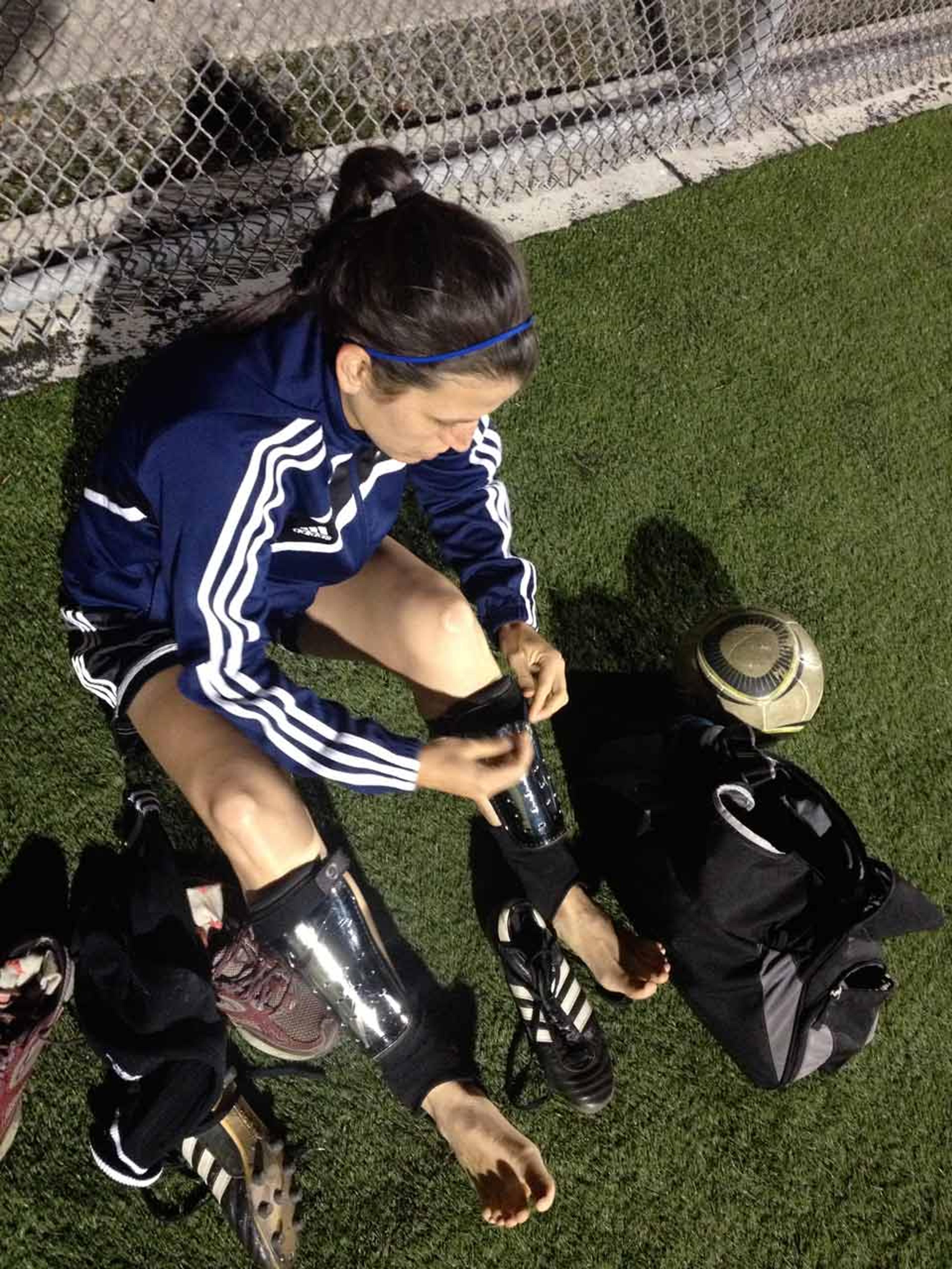 A female football player sitting on grass, putting on shin guards next to shoes and a bag by a chain-link fence.