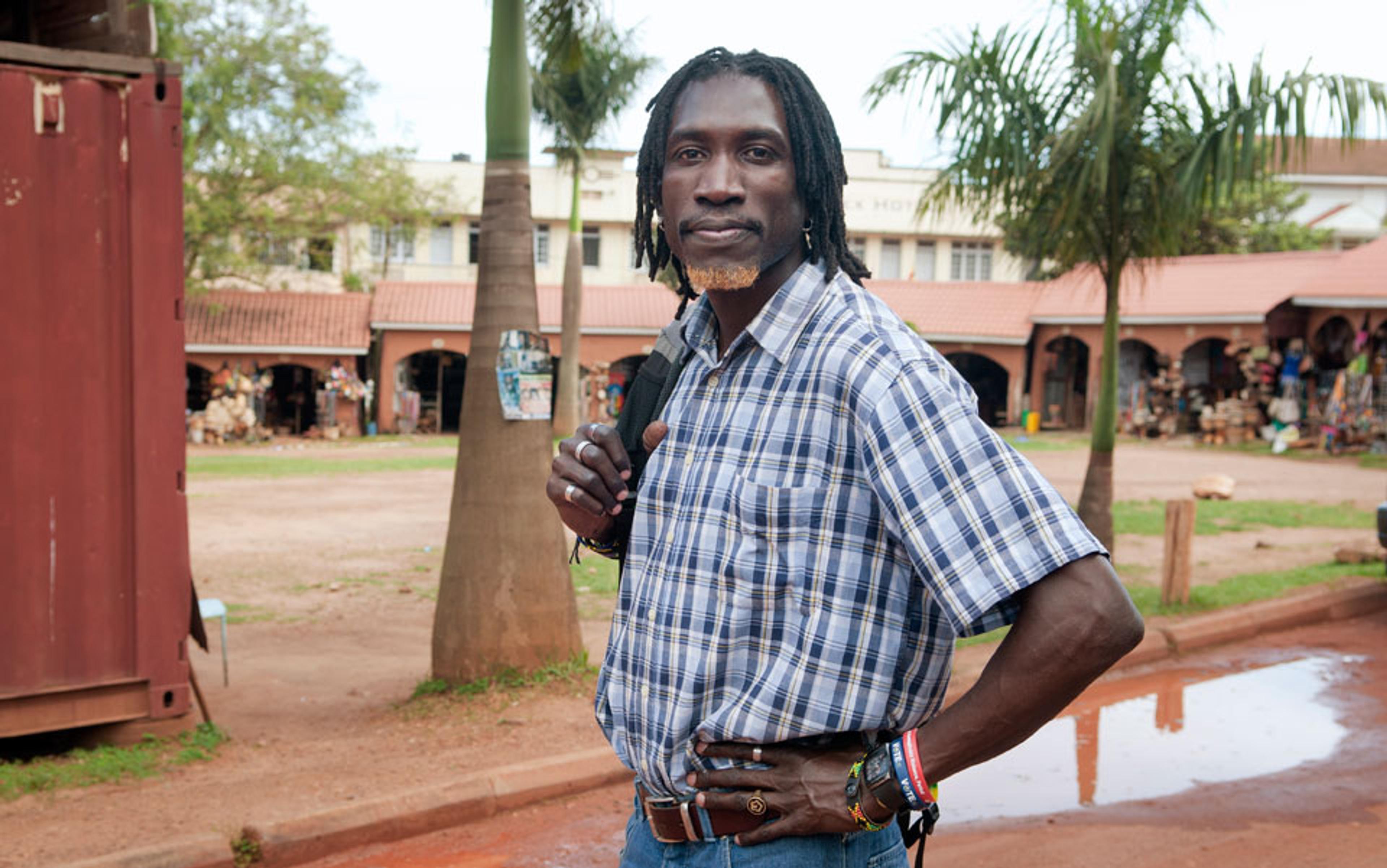 A Black man in a plaid shirt standing outside a marketplace with shops, wearing a backpack with palm trees in the background.