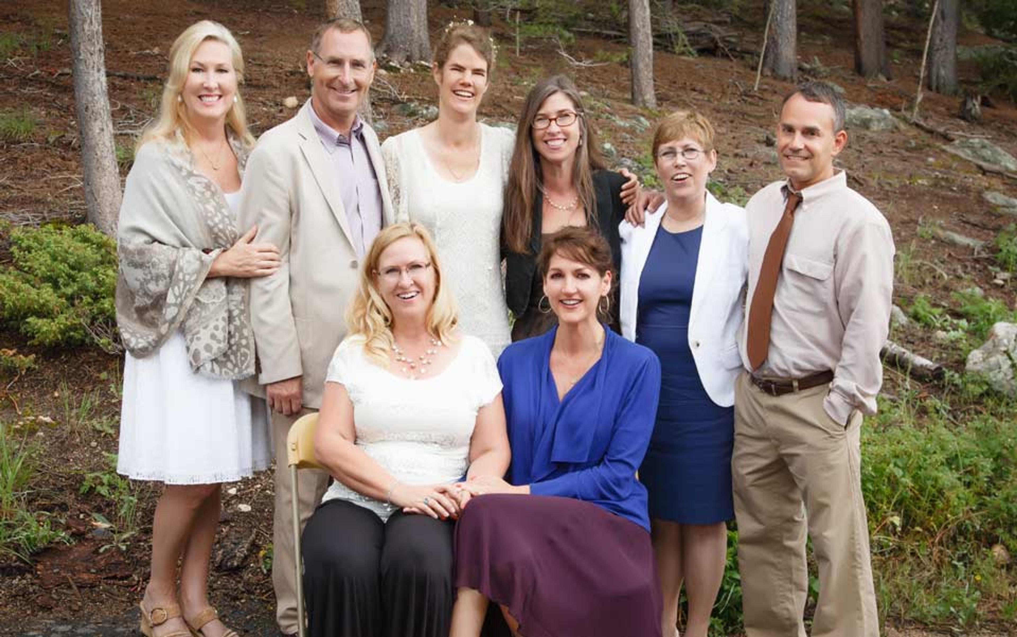 Photo of a group of eight adults, both seated and standing, posing outdoors in a wooded area, all smiling and dressed formally.