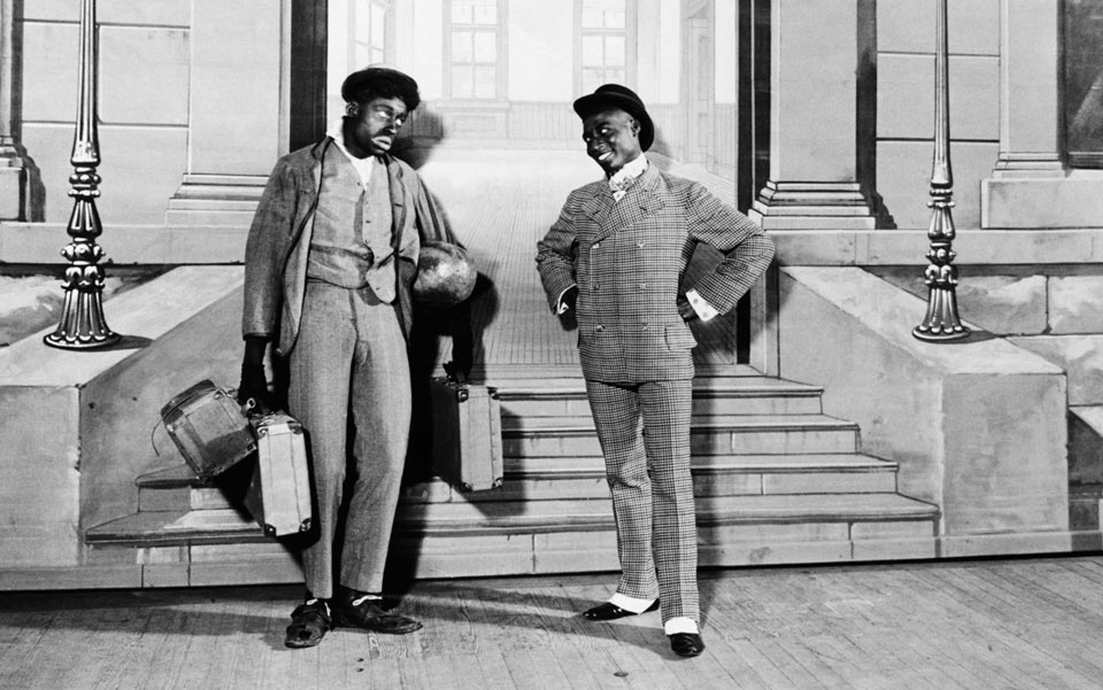 Black and white photo of two men in vintage attire on stage, one holding suitcases, in front of a painted backdrop of a building.