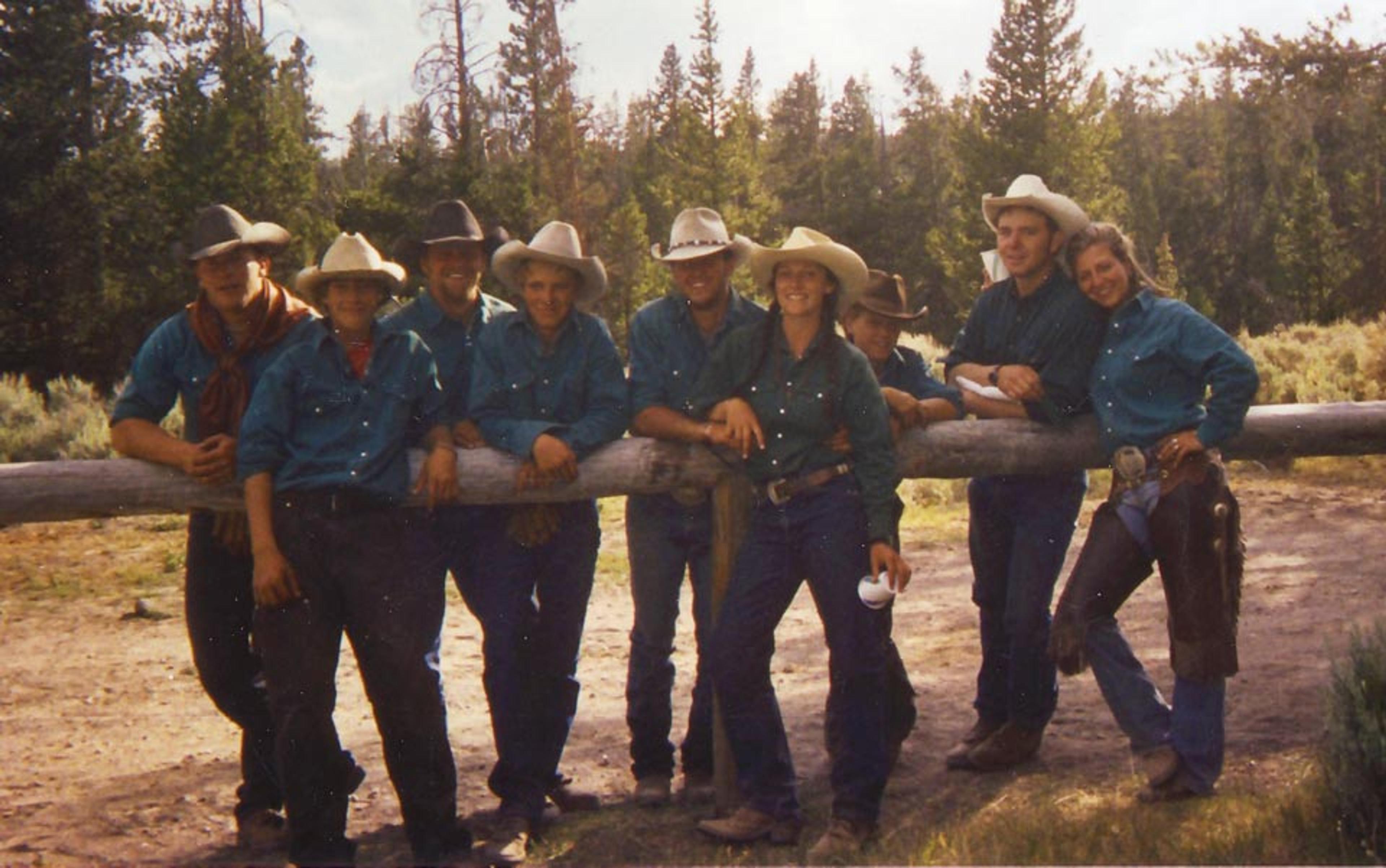 Eight people in cowboy attire standing and leaning against a wooden log with a forest background.