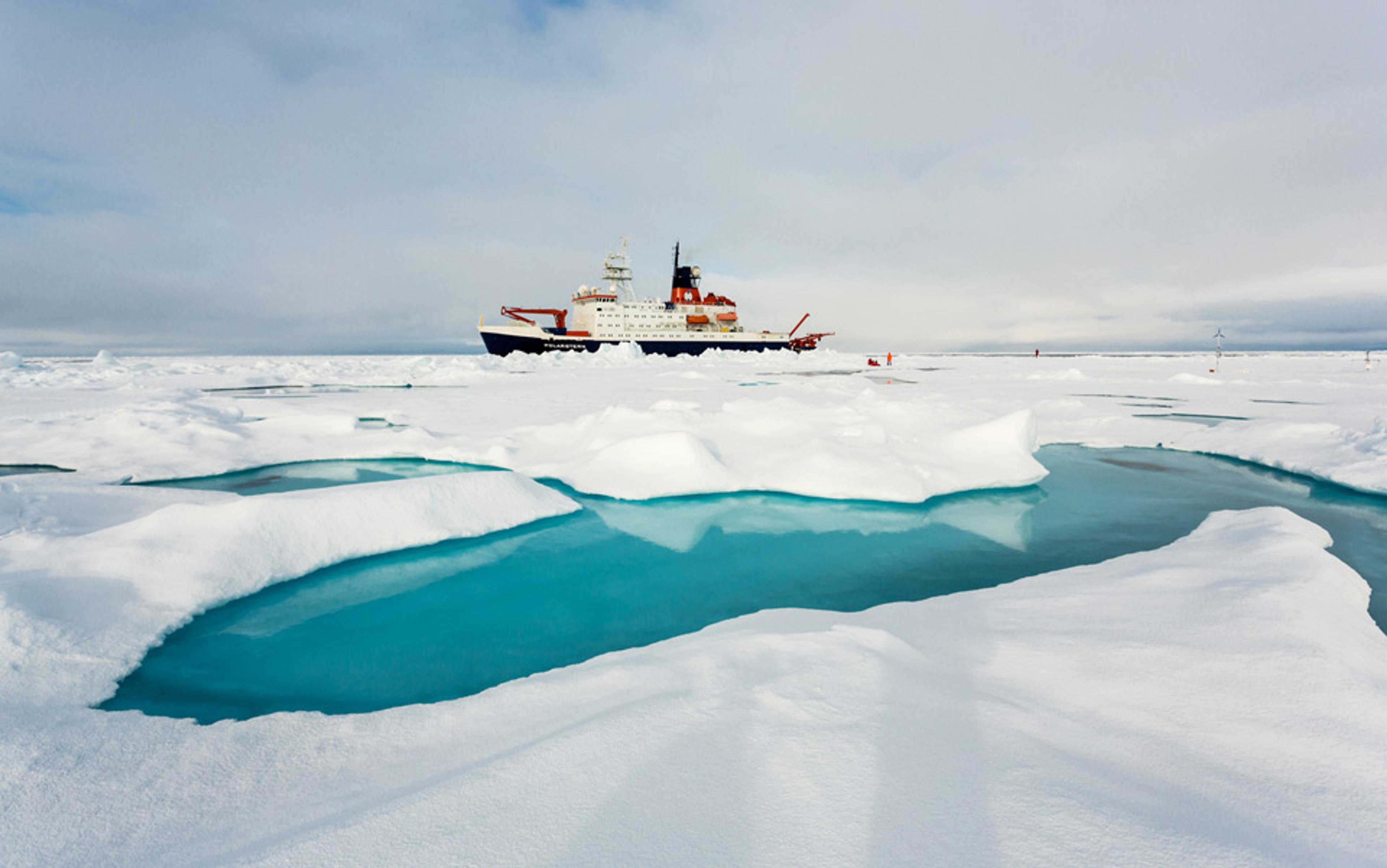 An icebreaker ship in the Arctic surrounded by ice and patches of blue water under a cloudy sky.