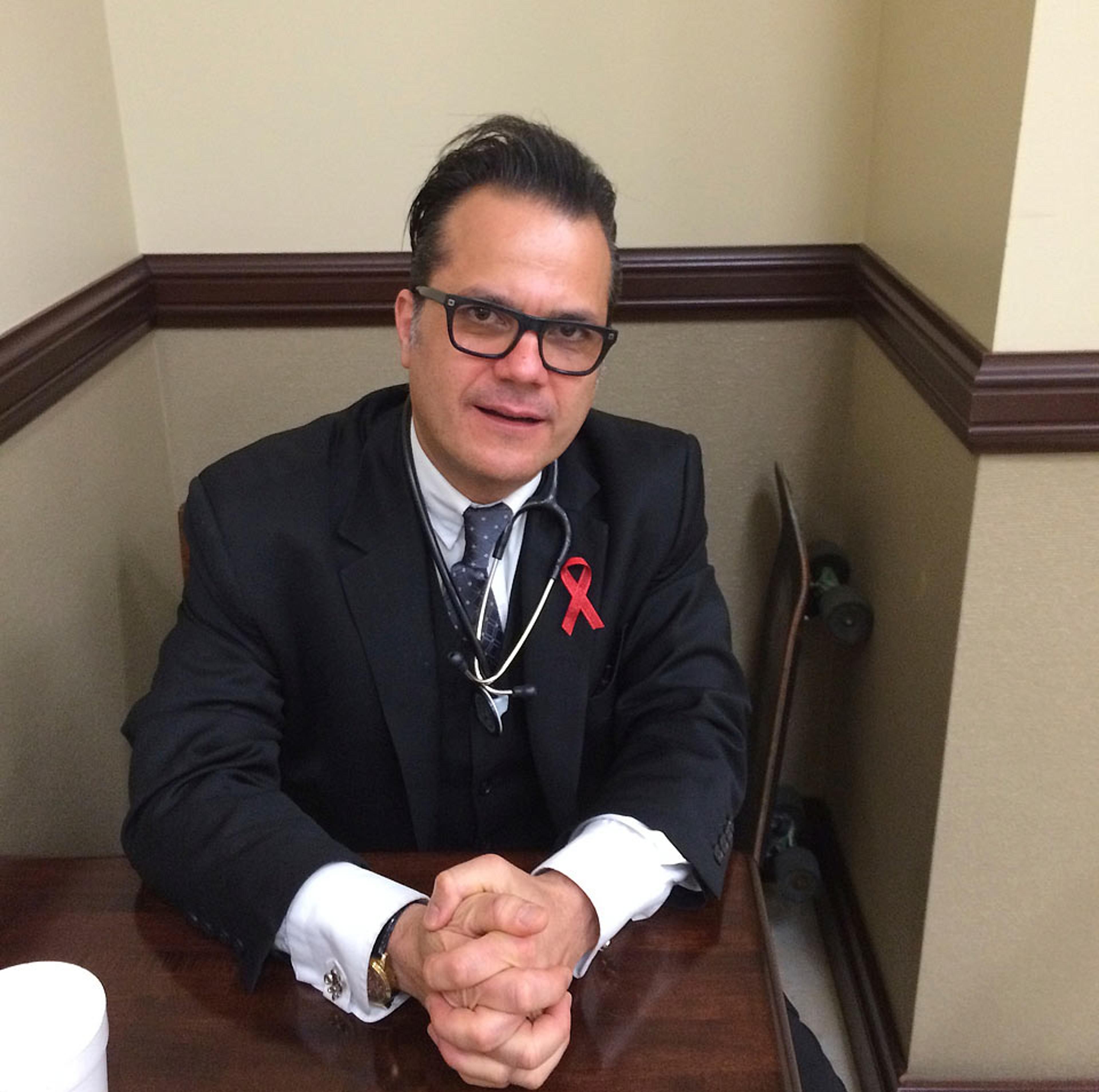 Photo of a man with glasses in a suit and stethoscope, sitting at a desk. He has clasped hands and a red ribbon on his lapel.