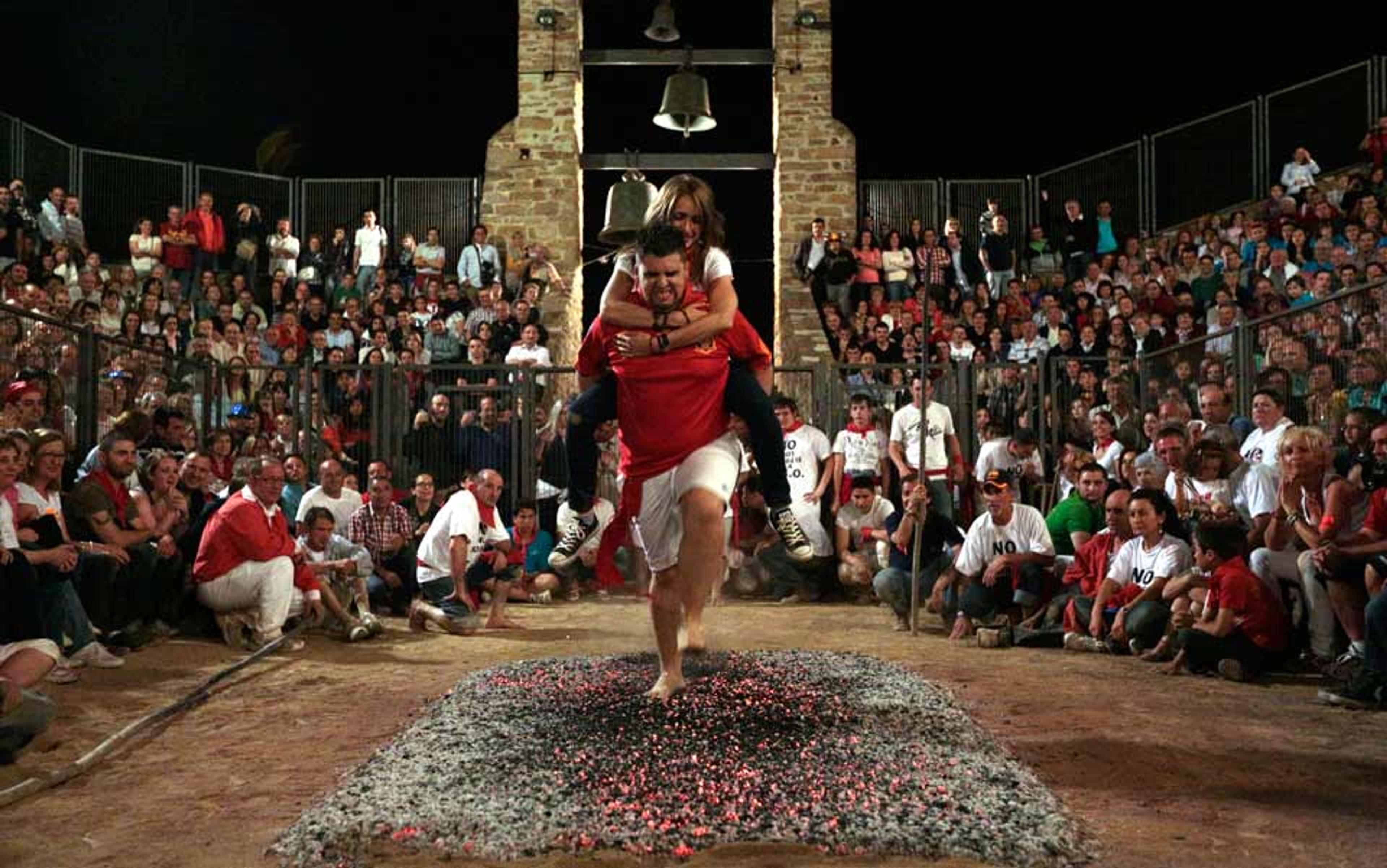 A man carrying a woman on his back as he walks barefoot over hot coals in an arena surrounded by a large crowd.