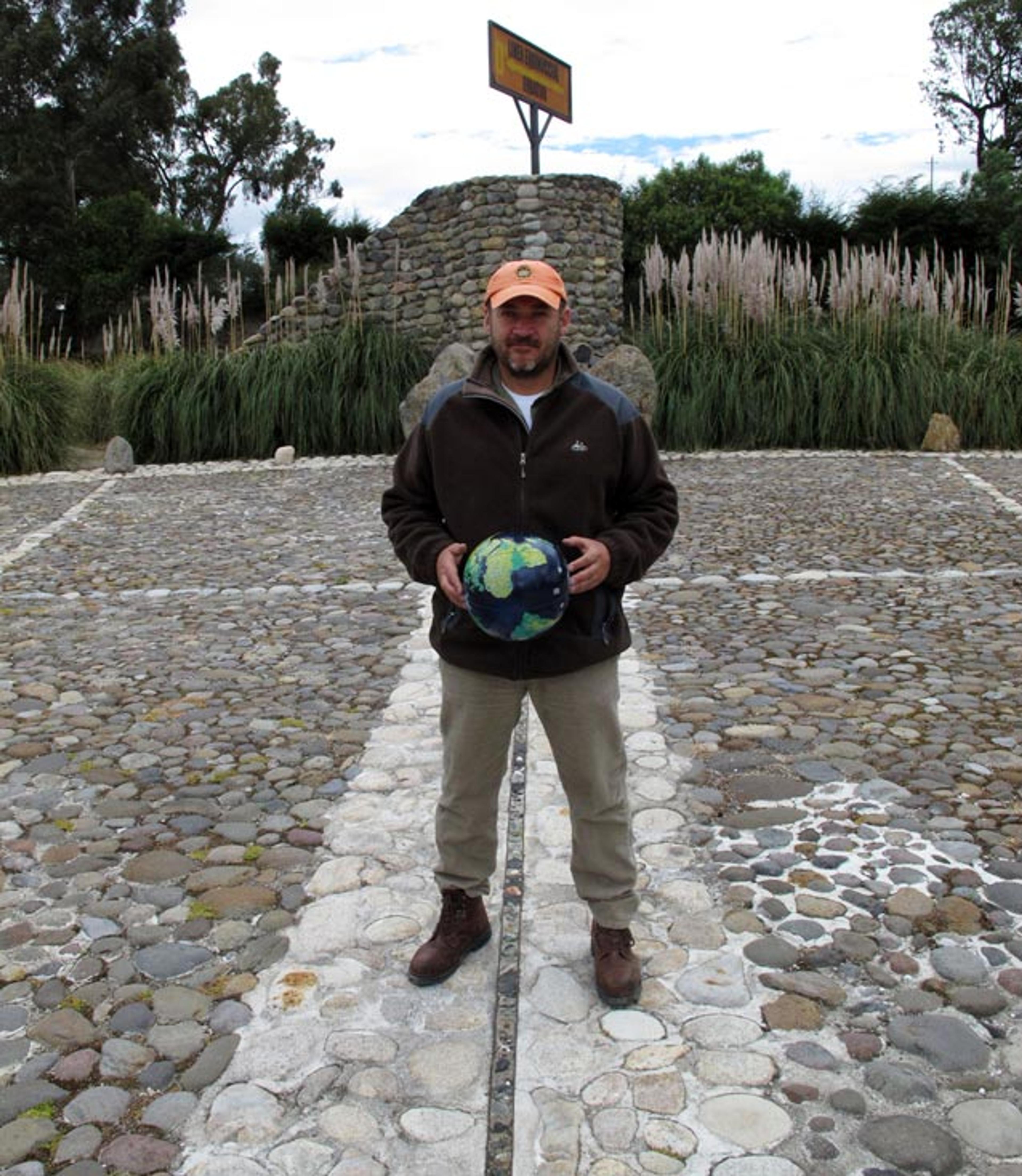 Photo of a man standing on a cobblestone pathway holding a globe, with greenery in the background and a stone structure behind him.