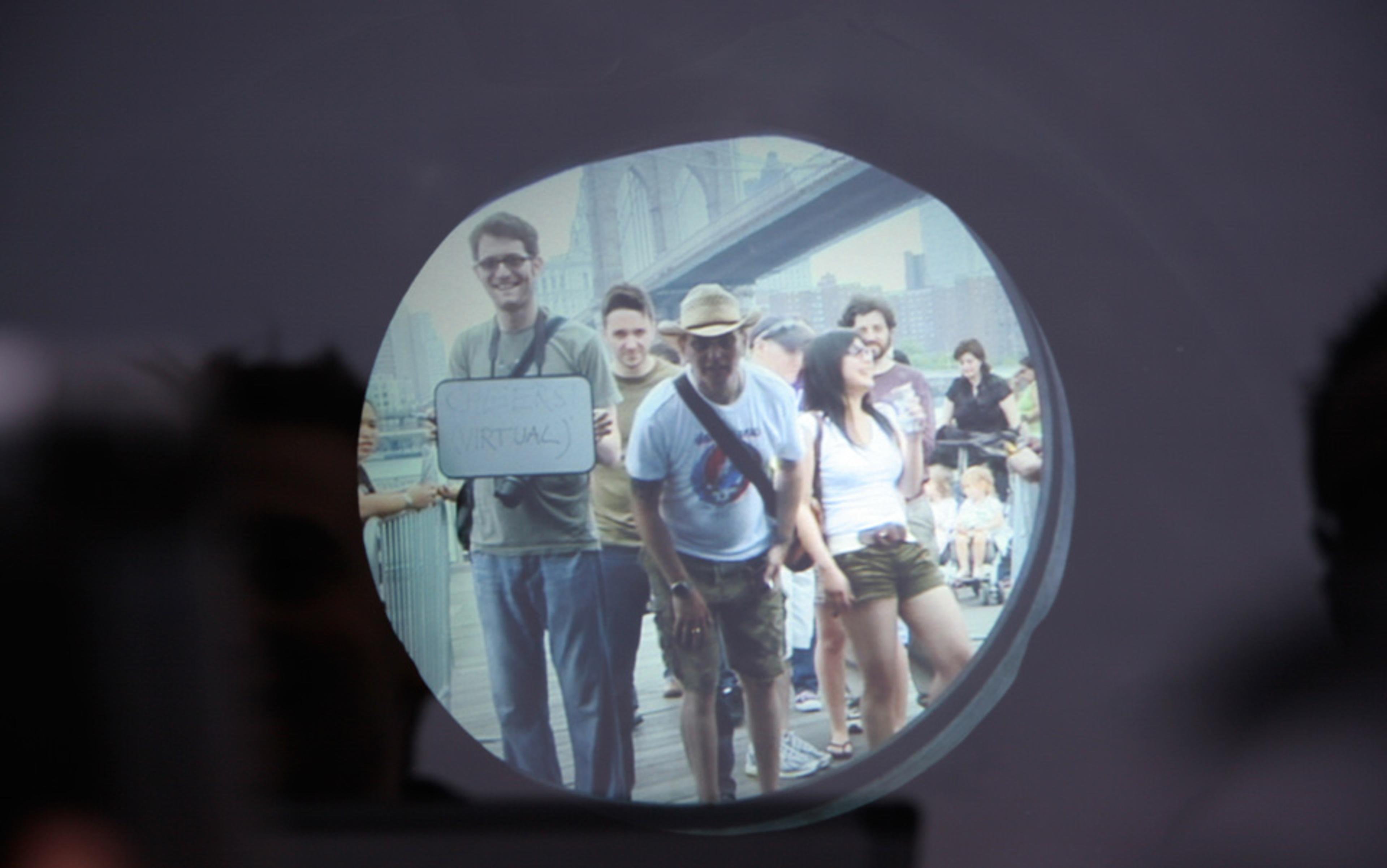 Photo of a group of people viewed through a circular opening with a blurry reflection around the edge. A bridge is visible in the background.