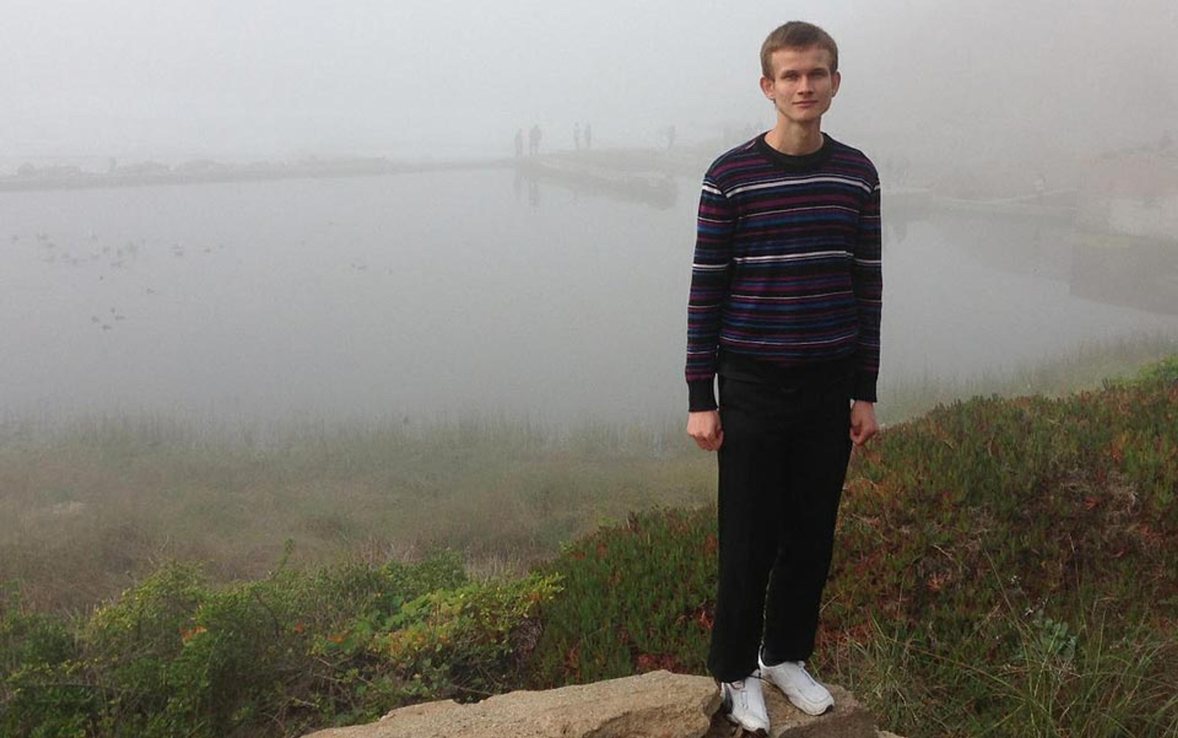 A man in a striped sweater standing on a foggy lakeside with greenery and faint figures visible in the background.