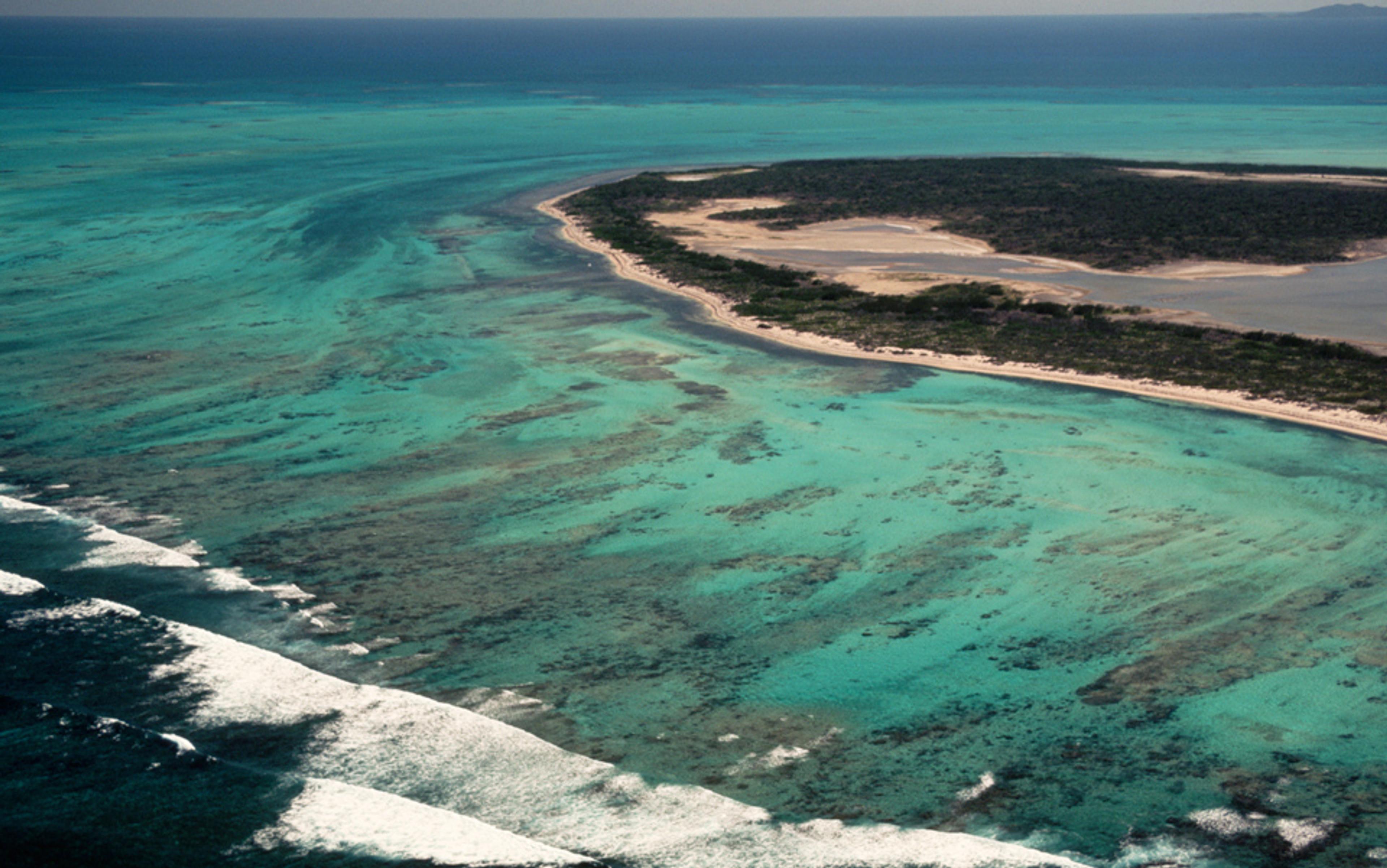 Aerial photo of a coastal area with a turquoise sea, a sandy shoreline, and vegetated land, with waves breaking near the shore.