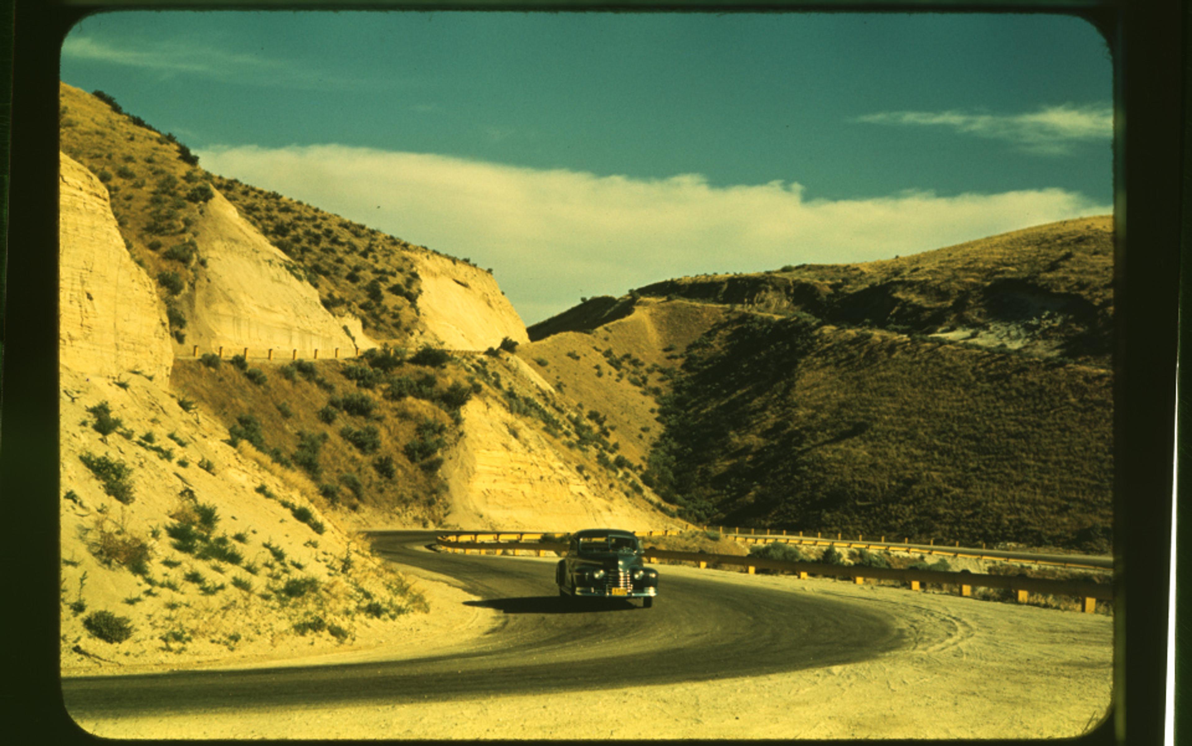 A vintage car on a winding road through arid hills under a clear sky, photo from the mid-20th century.