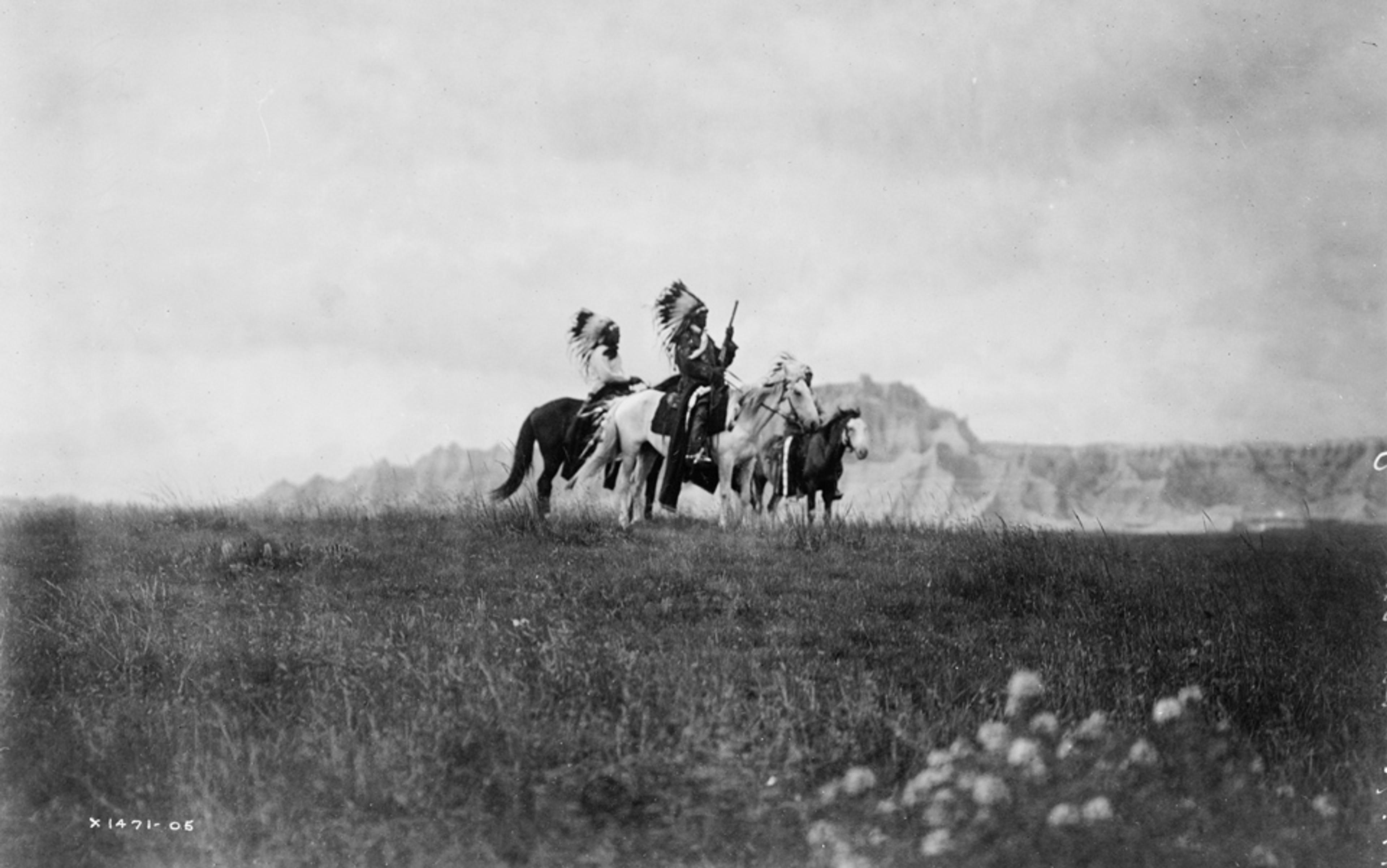 Black and white photo of two people wearing Native American attire riding horses on a grassy plain with mountains in the background.