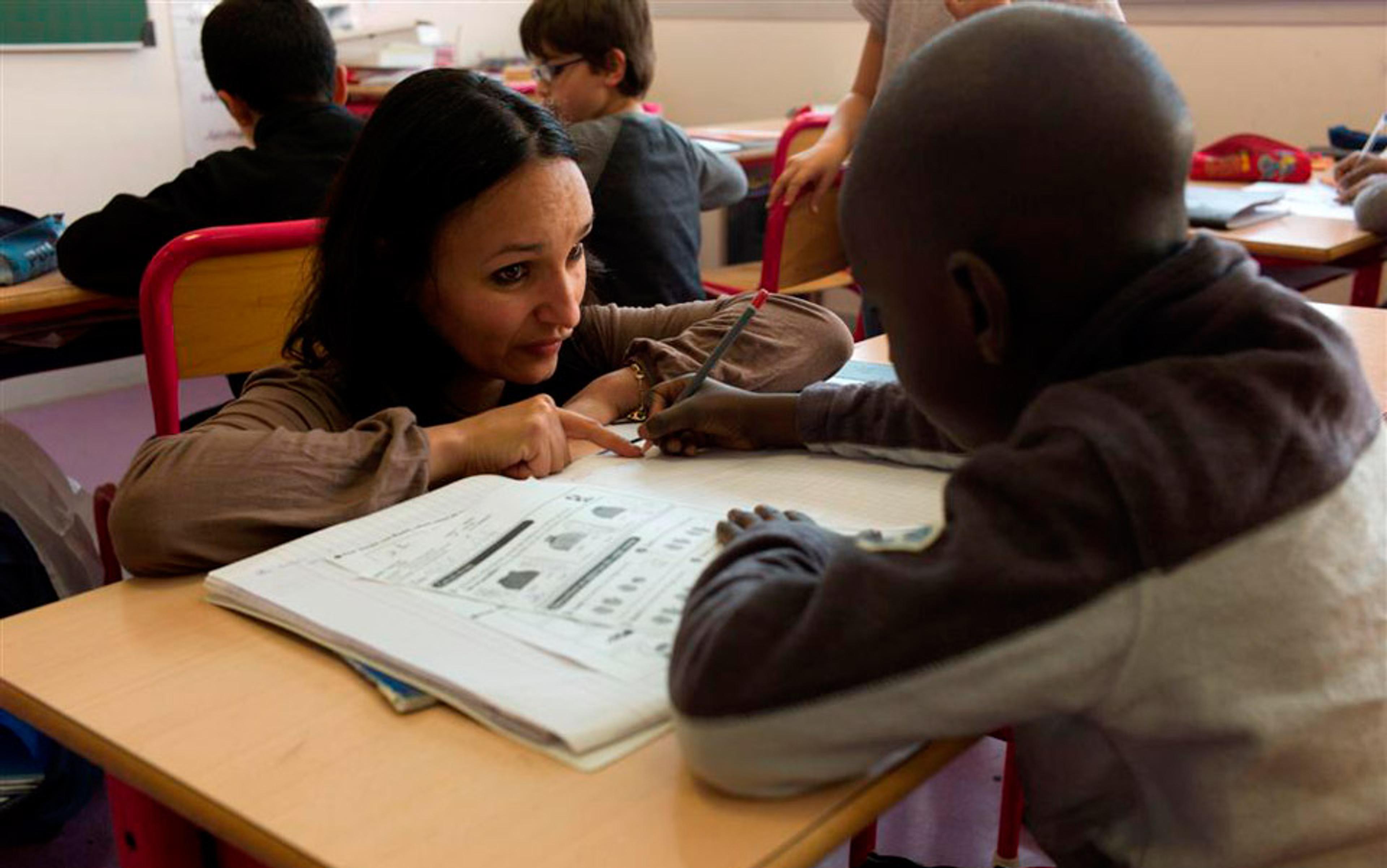 Photo of a teacher assisting a student with a workbook in a classroom, surrounded by other students working at their desks.