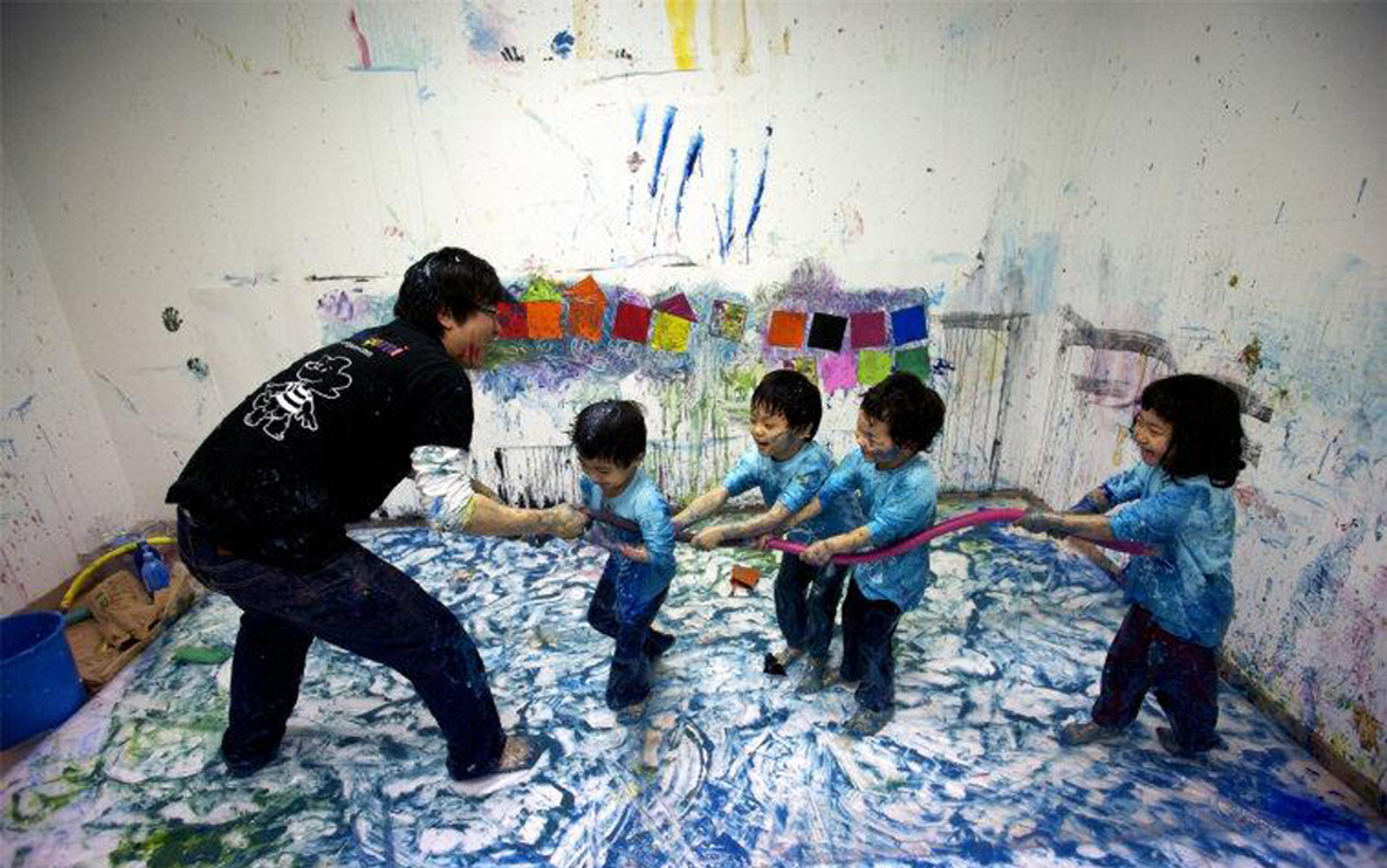 Photo of an adult and four children in a messy art room pulling on a rope, with colourful paintings on the walls and floor.