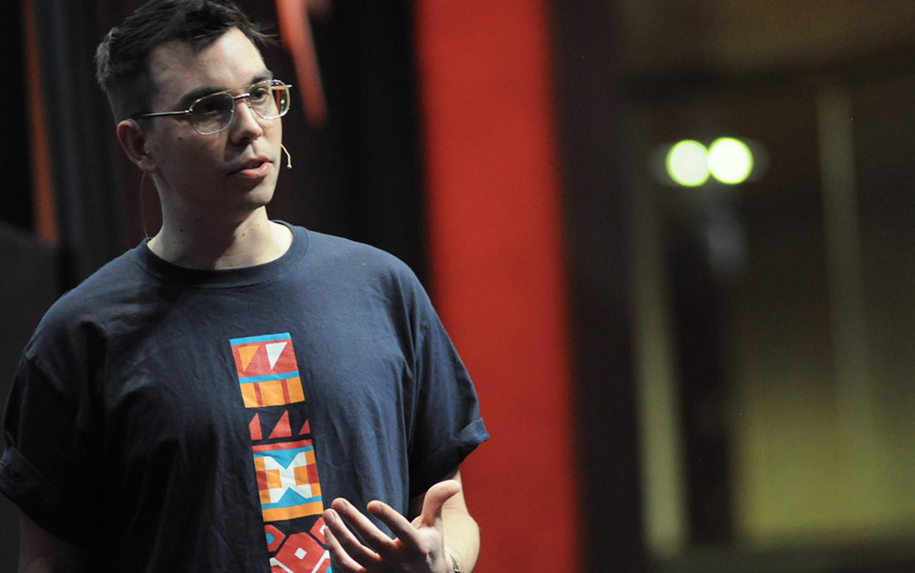 A person wearing glasses and a T-shirt with a colourful pattern, speaking into a headset microphone on a stage.