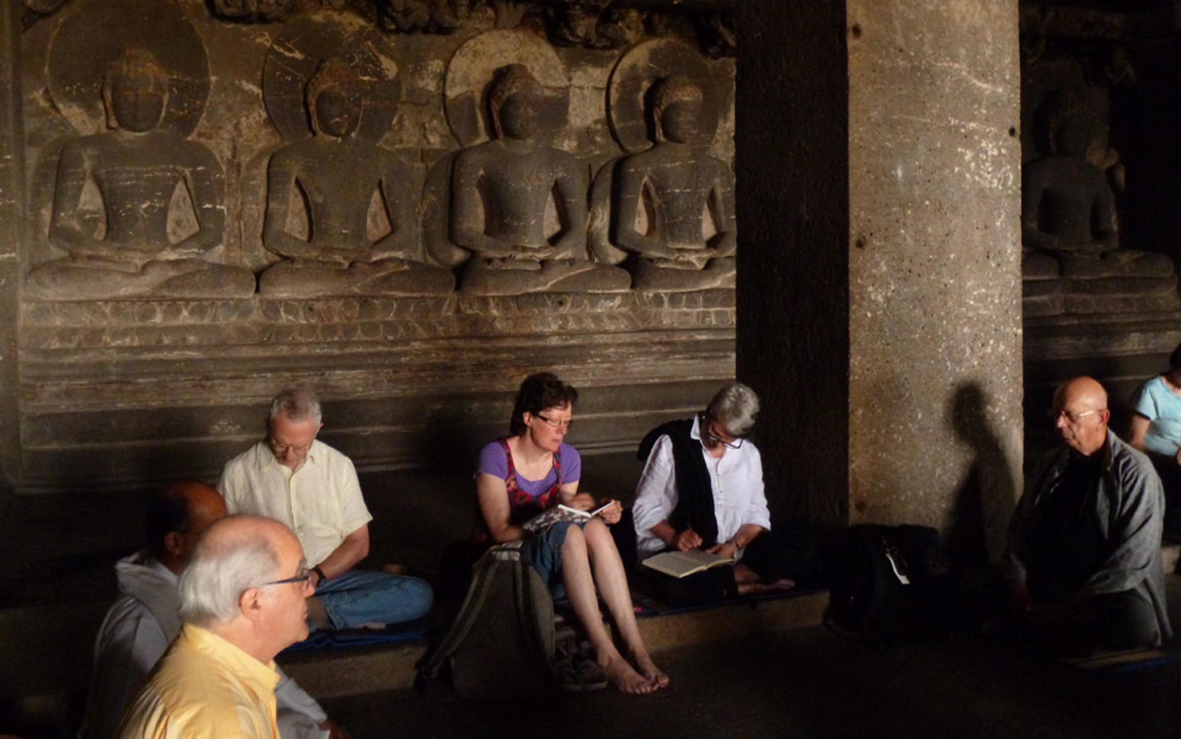 A photo of people meditating and writing in a stone temple with seated Buddha statues carved into the wall behind them.