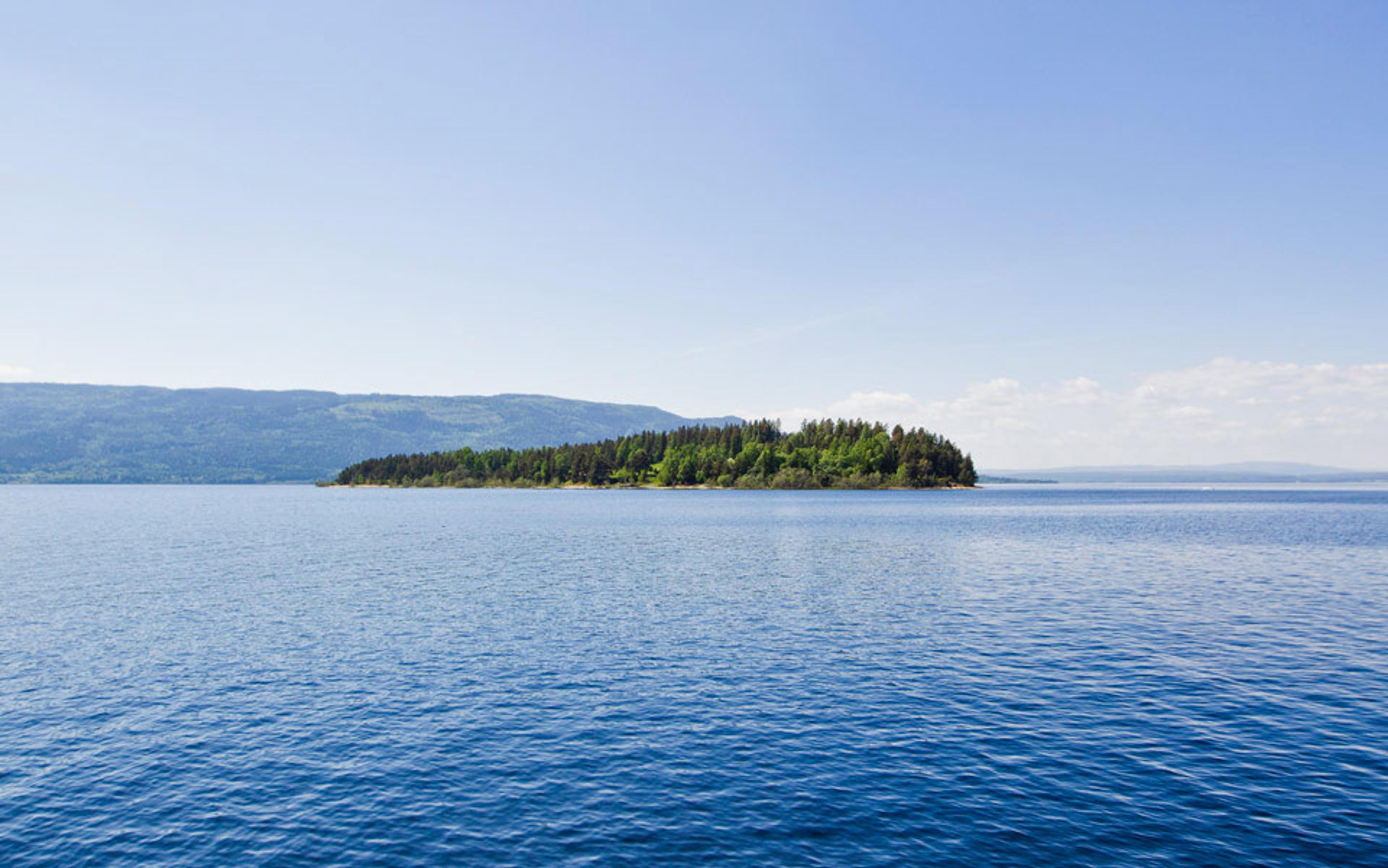 Photo of a small, forested island surrounded by calm blue water with distant mountains and a clear sky in the background.