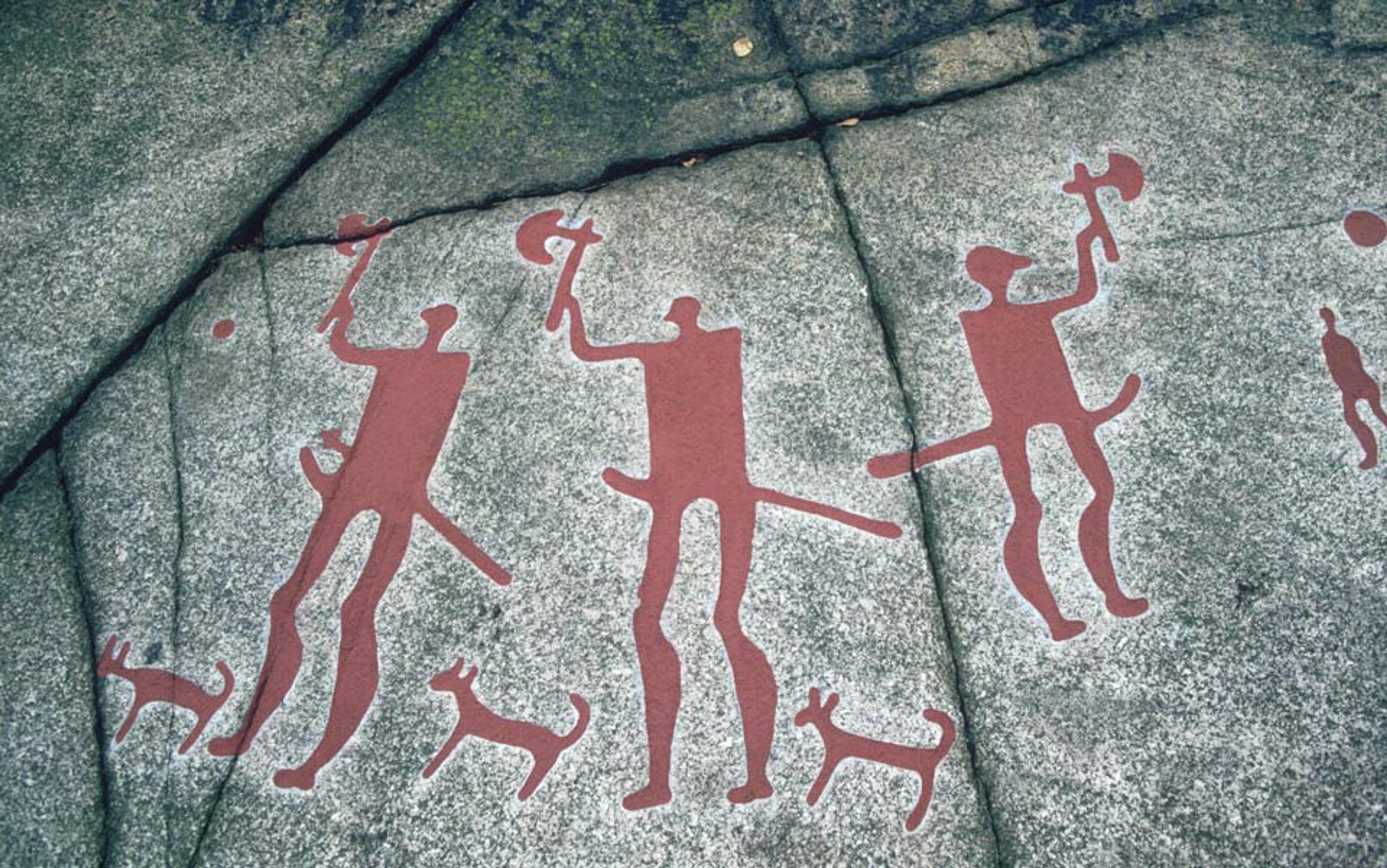 Ancient rock painting depicting red figures of people and dogs holding tools on a grey stone surface in a prehistoric style.