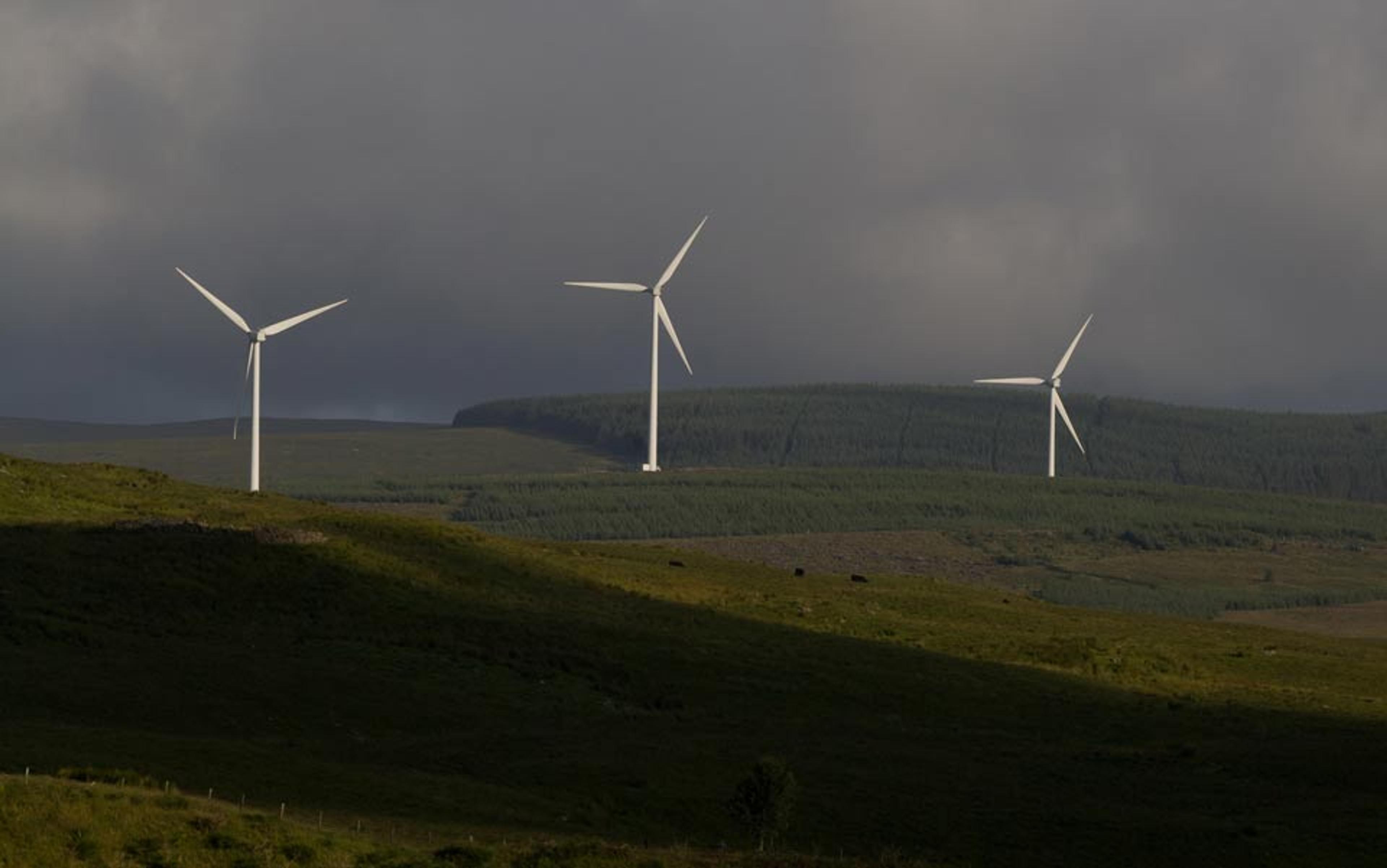 Three wind turbines on a grassy hillside with a forested background and dark, stormy clouds in the sky.