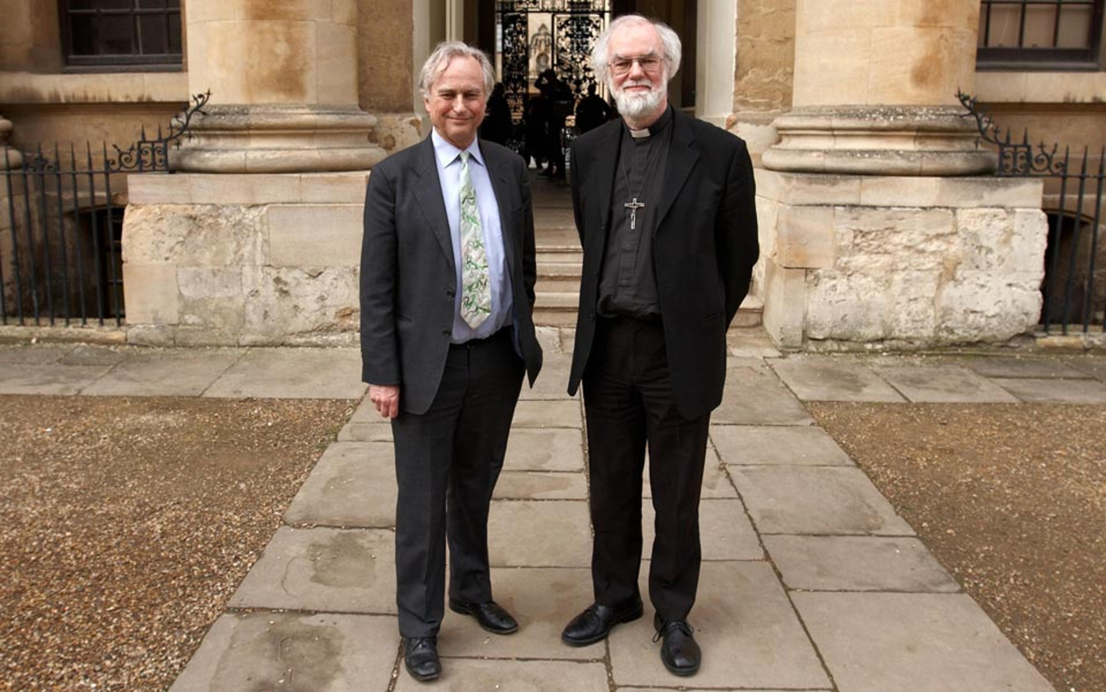 Photo of two men standing in front of a historic stone building, one in a suit with a patterned tie, the other in clerical attire with a cross.