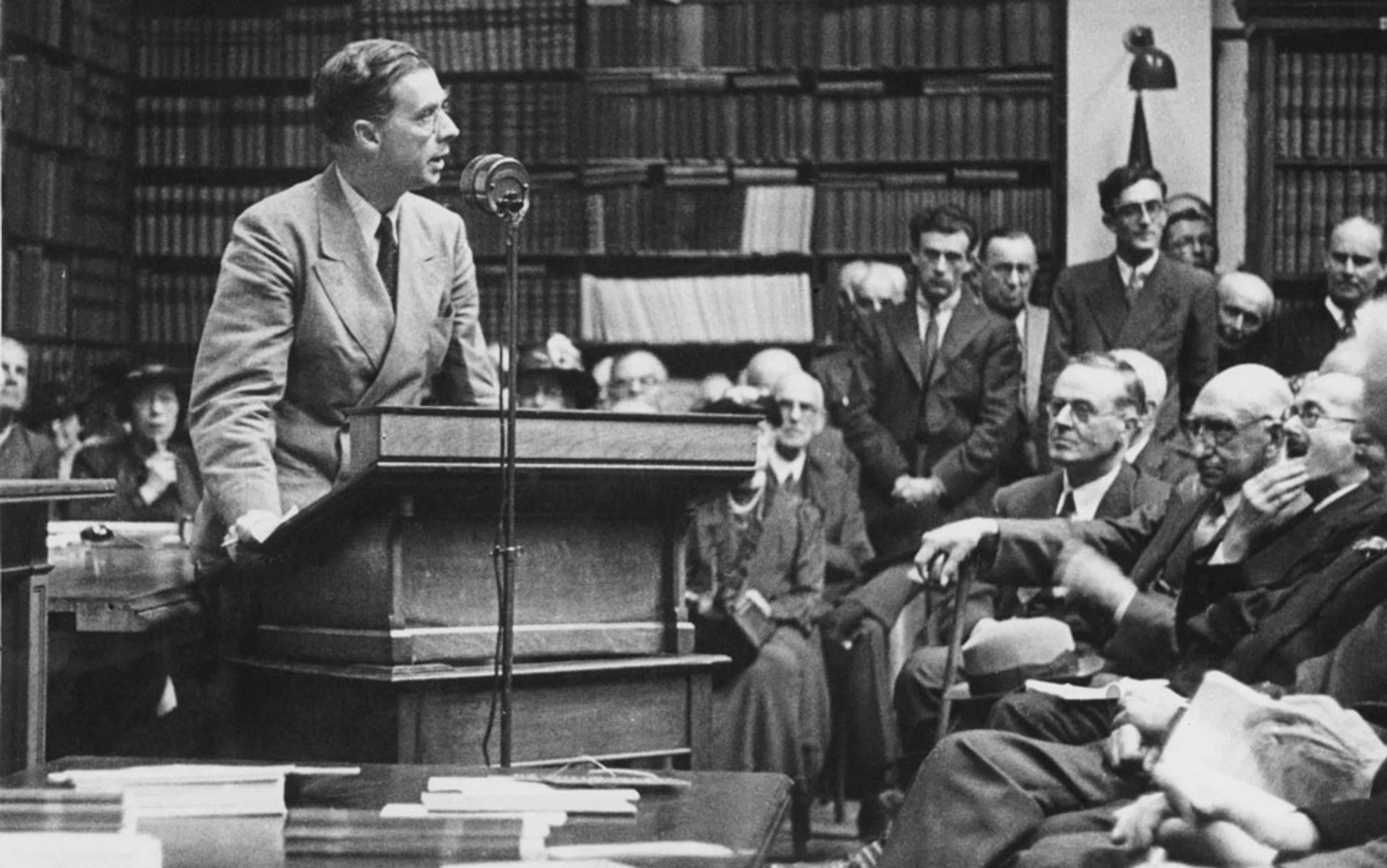 Black-and-white photo of a man giving a speech at a podium to an audience in a library setting with bookshelves in the background.