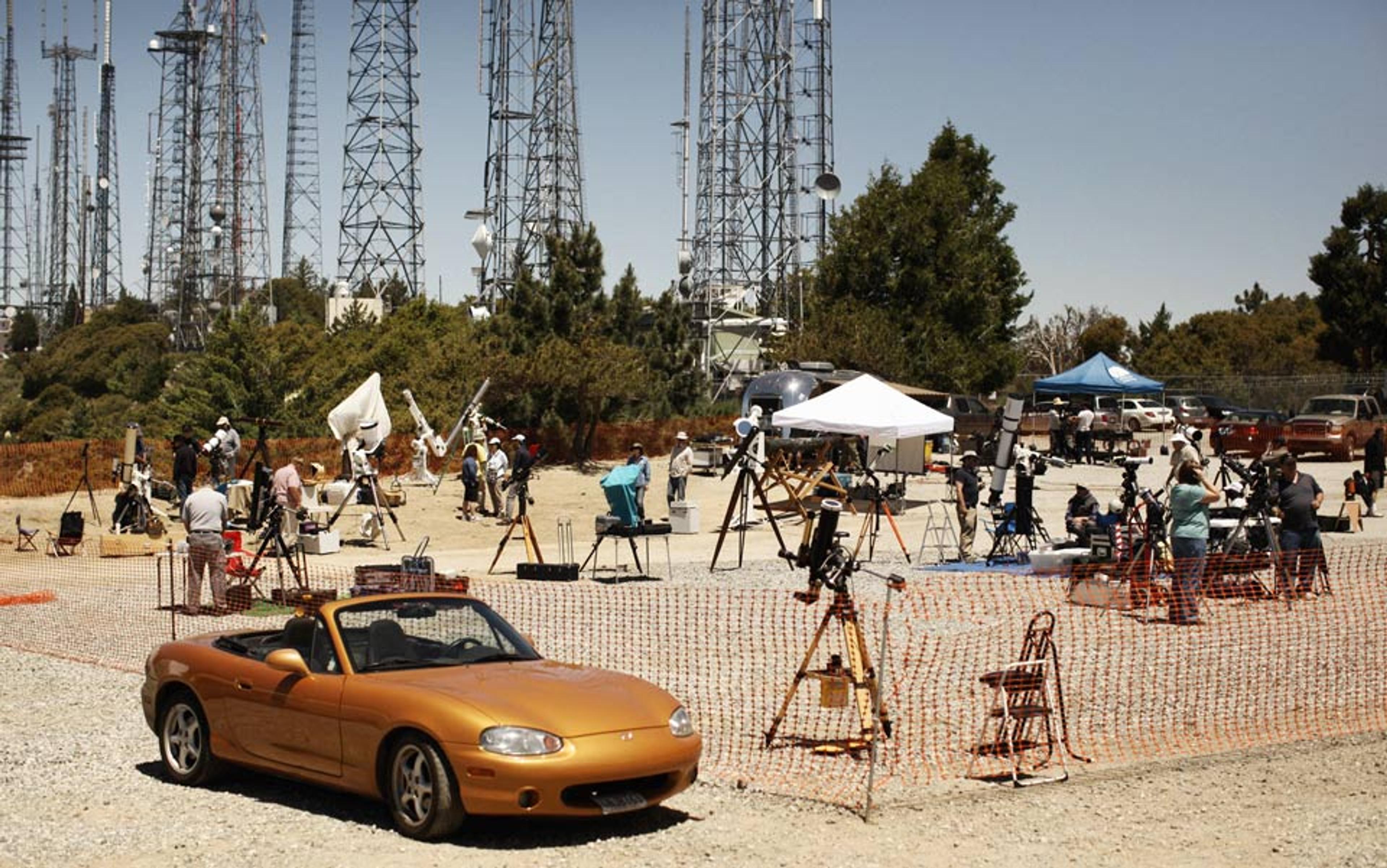 Photo of a gathering of amateur astronomers with telescopes and equipment, set up near numerous radio towers and a gold convertible car.