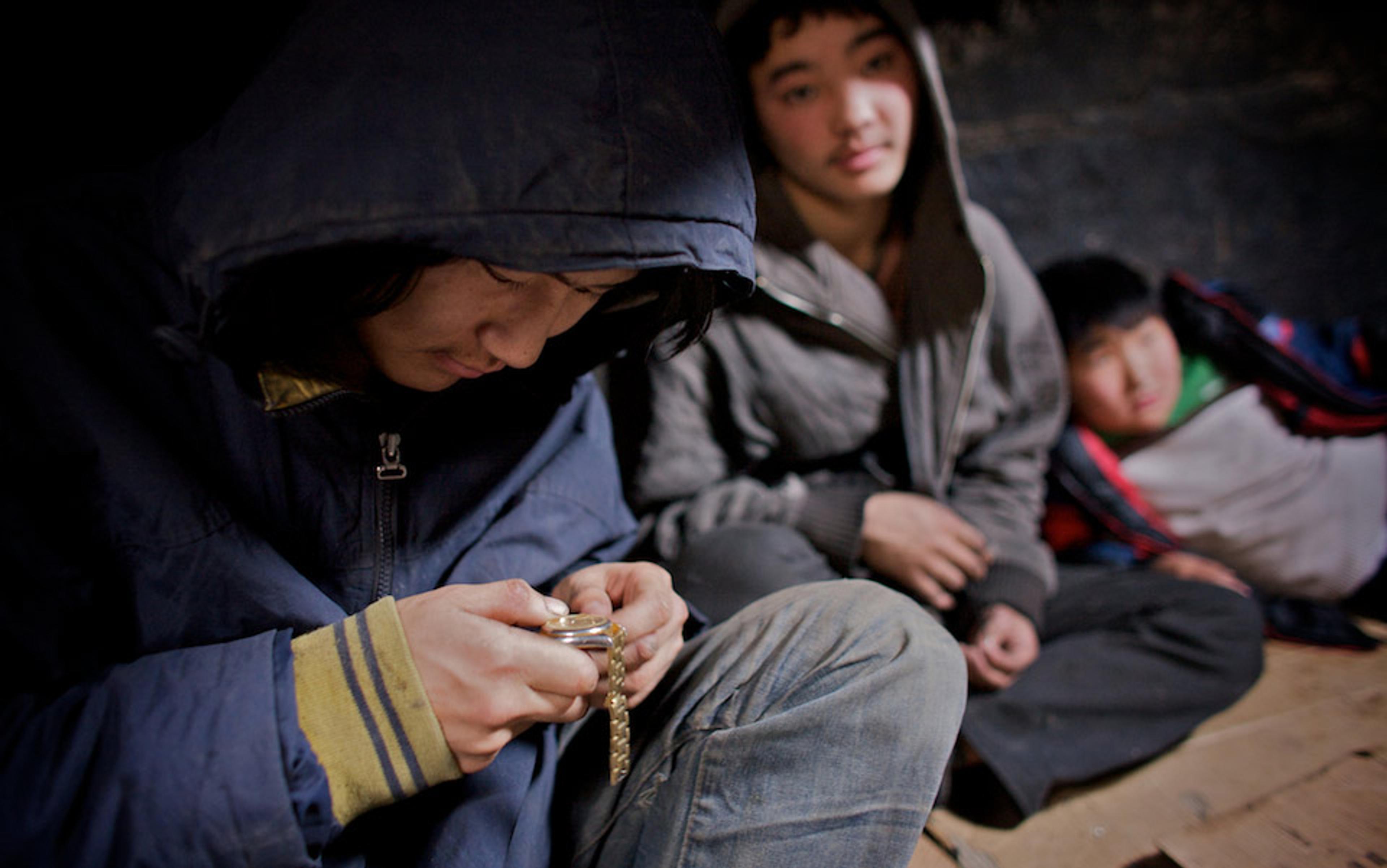 Three Mongolian boys indoors wearing hoodies, with one focused on a gold watch in his hands while the others sit observing in the background.