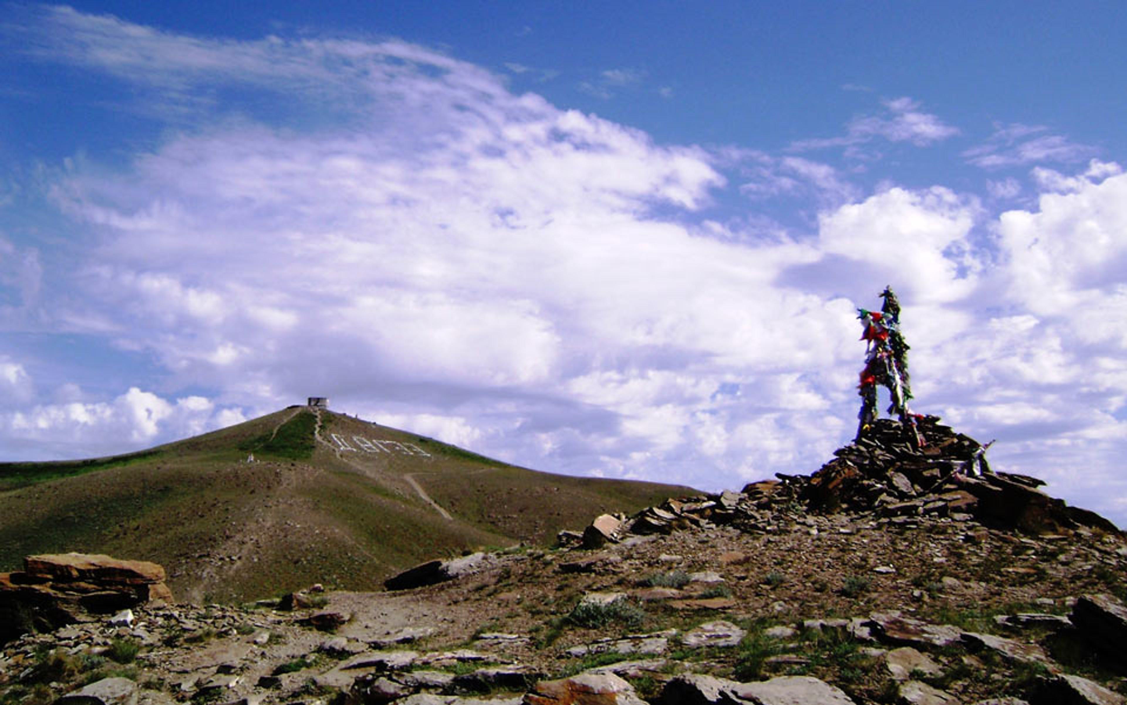 A rocky hill with a small structure on top and prayer flags on a stone pile in the foreground, under a partly cloudy sky.