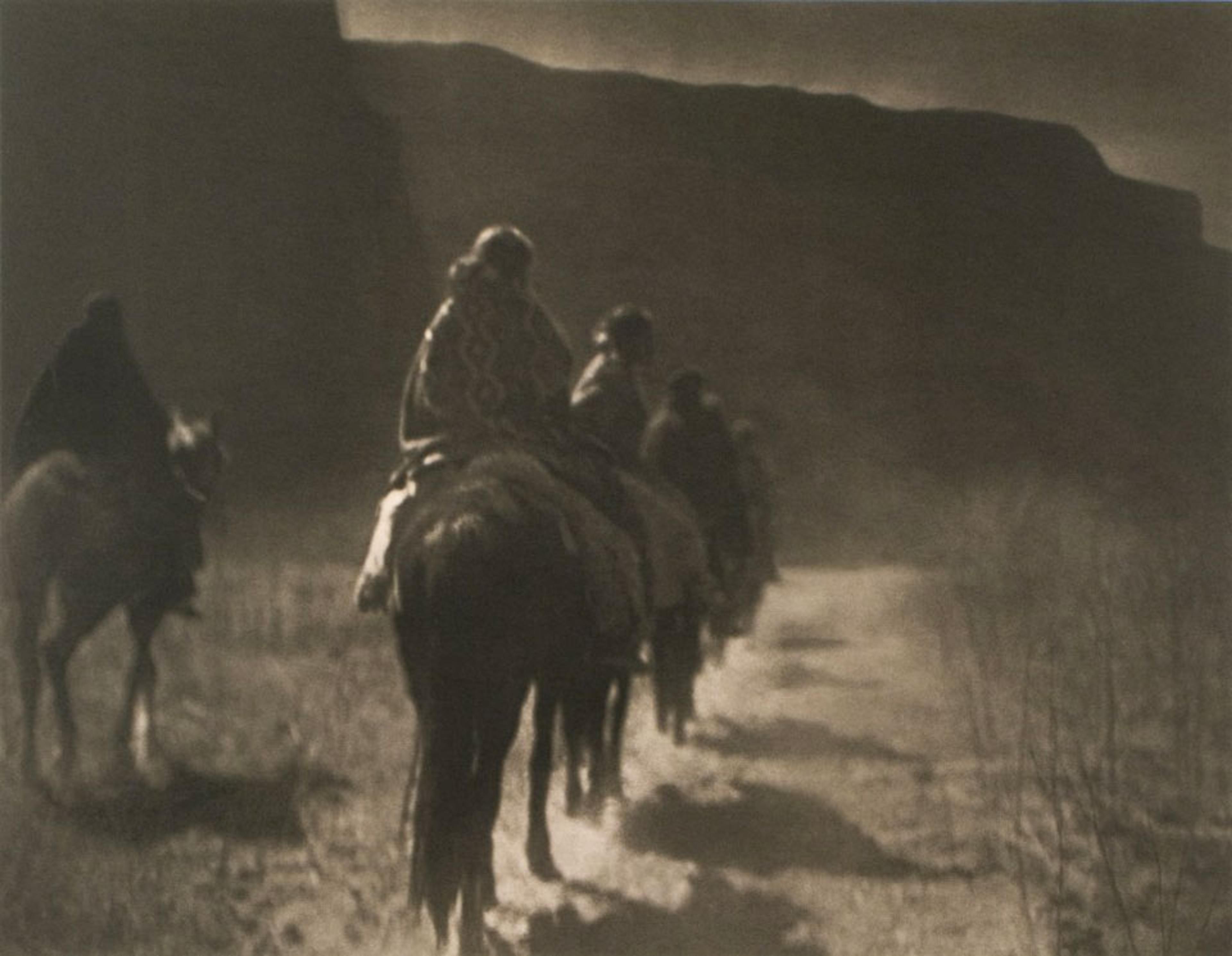 Sepia-toned photo of several people riding horses in a line along a dirt path, with cliffs and shadows in the background.