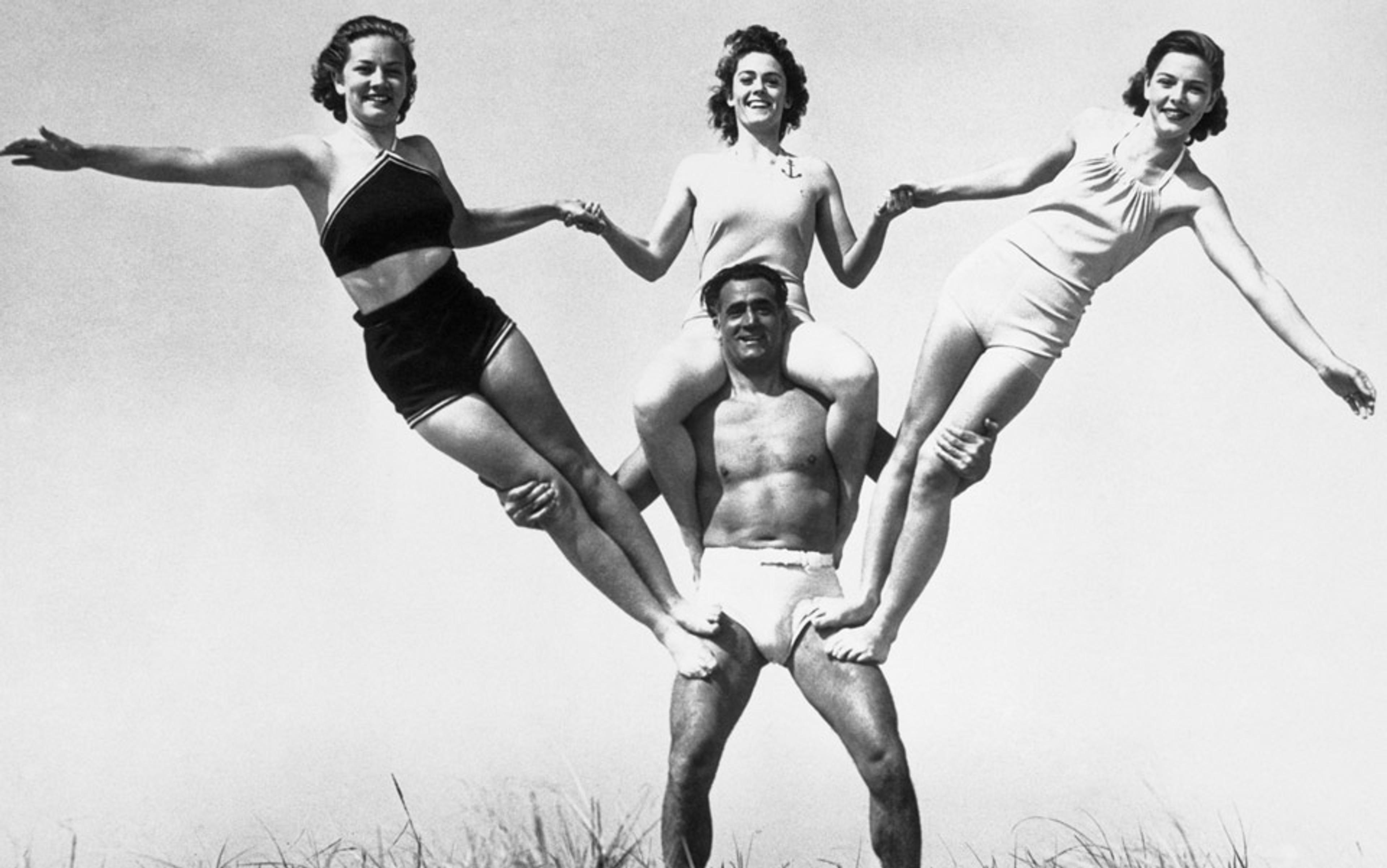 Black and white photo of a man balancing three women in swimsuits, two standing on his shoulders and one on his arm, against a clear sky.