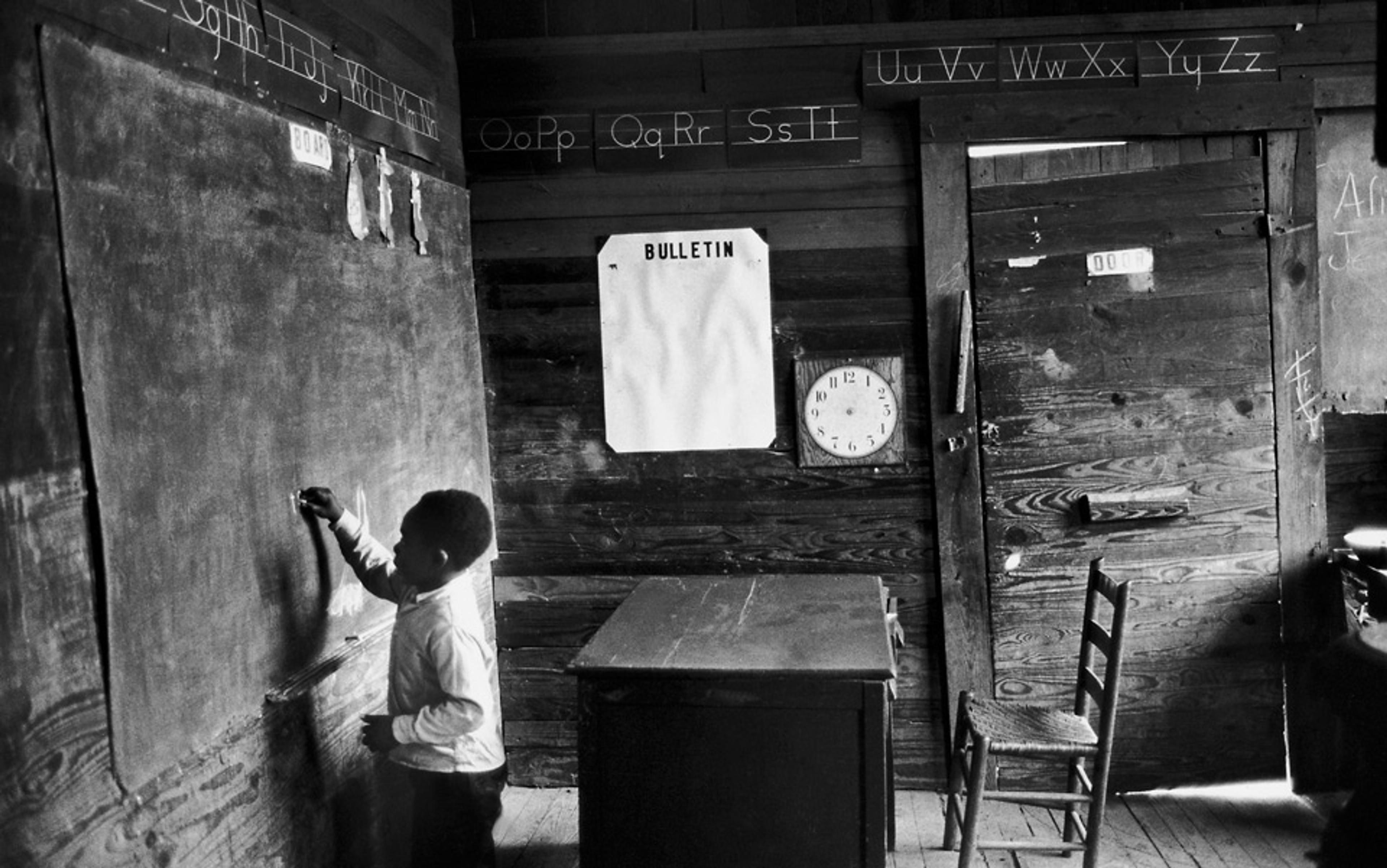 Black-and-white photo of a Black child writing on a chalkboard in a classroom with an empty bulletin board, alphabet letters, a clock and chairs.