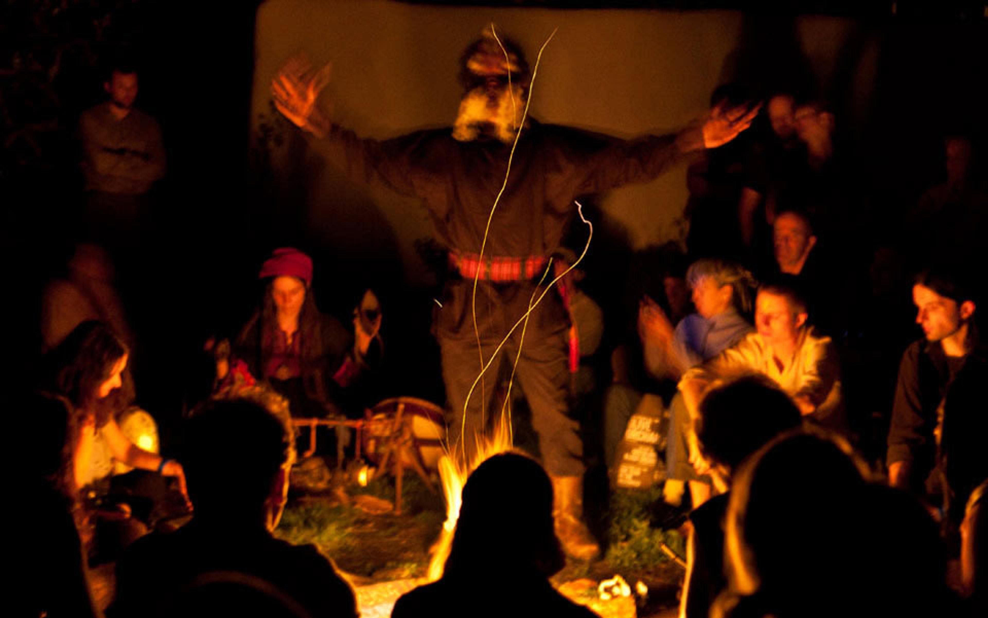 A photo of a figure with arms raised in the centre of a circle of people sitting around a bonfire at night.