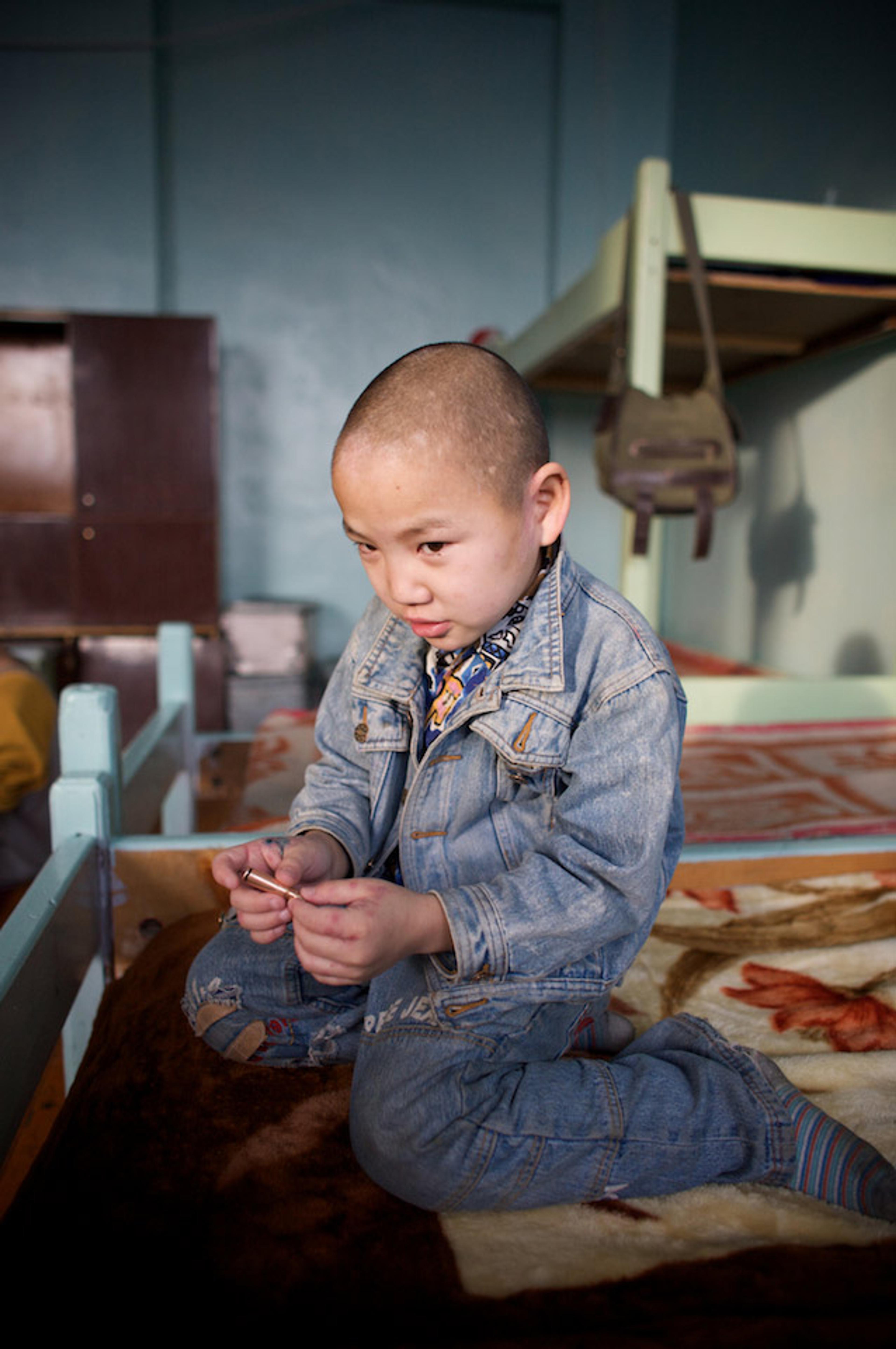 A young Mongolian boy in a denim jacket kneeling on a bed in a room with bunk beds, holding a small object and looking down.