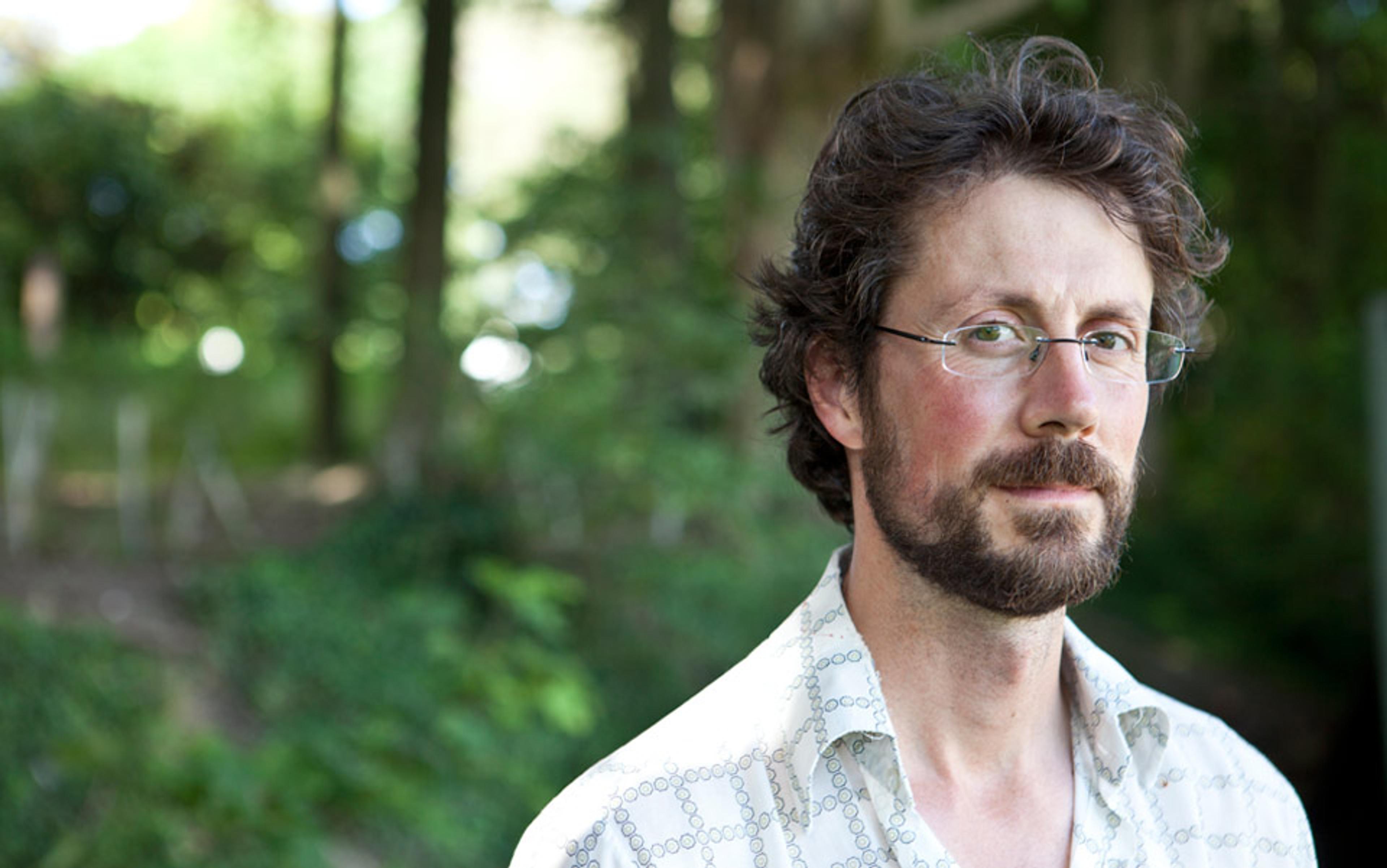 Photo of a man with glasses and a beard wearing a patterned shirt standing outdoors with a blurred green forest background.