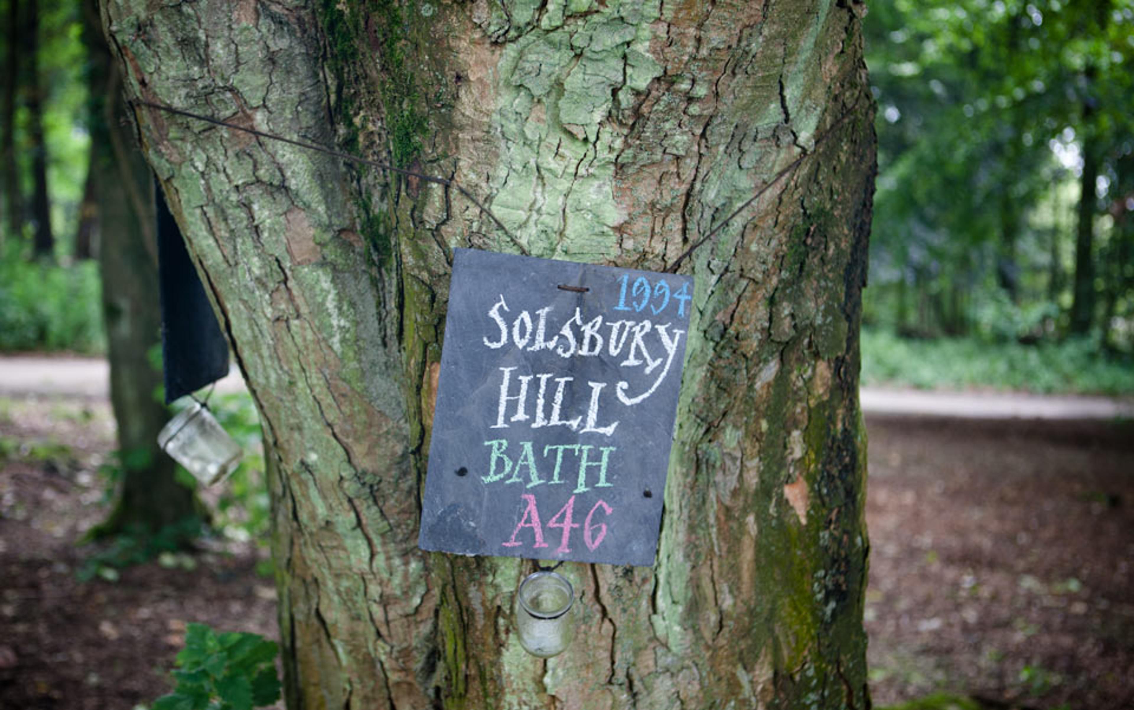 A photo of a tree with a handwritten chalk sign that reads “Solsbury Hill, Bath, A46, 1994” in a forested area.