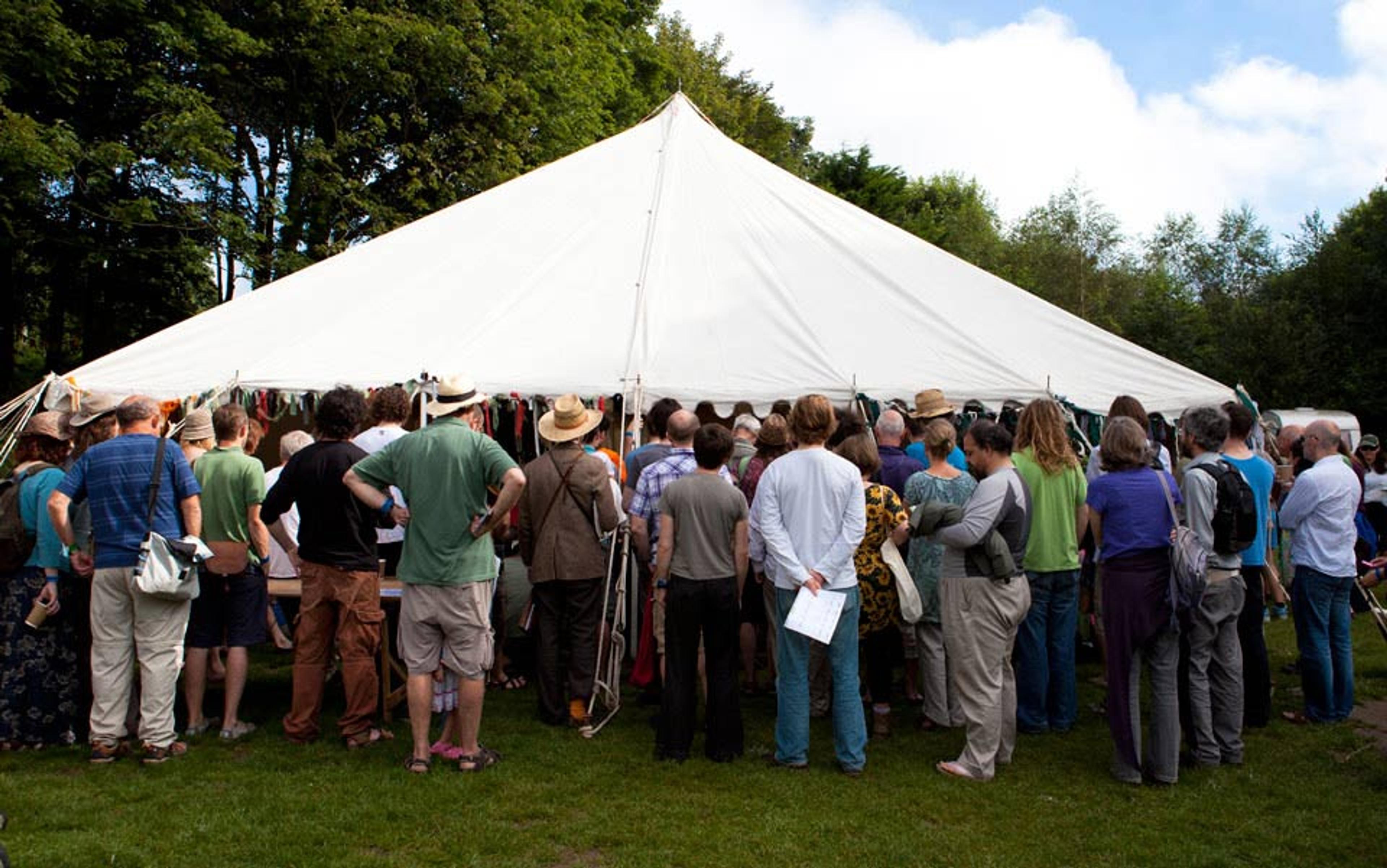 Photo of a crowd of people gathered around a large white tent in a grassy outdoor area with trees in the background.