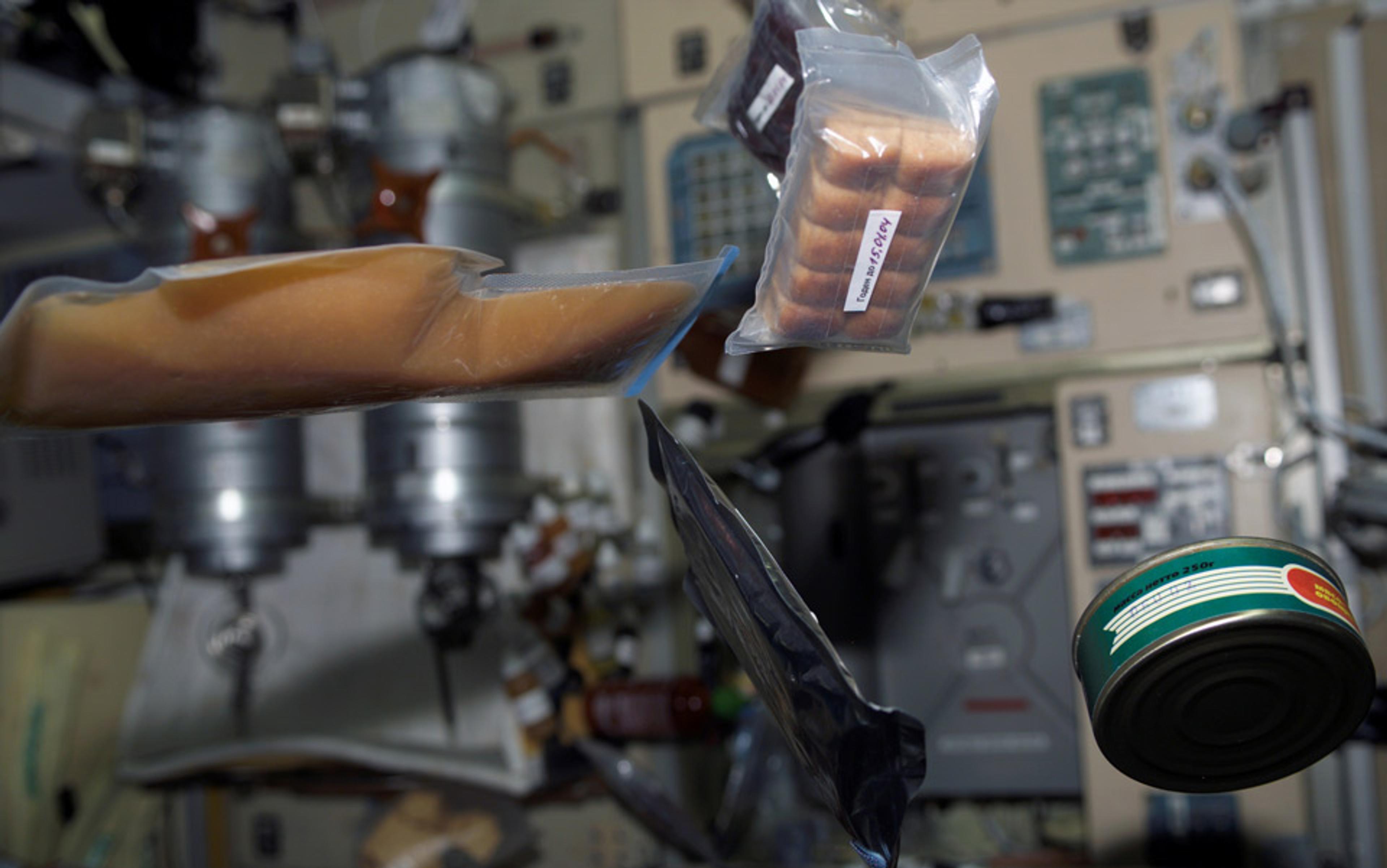 Floating packaged food and a tin can inside a spacecraft, with control panels and equipment in the background.