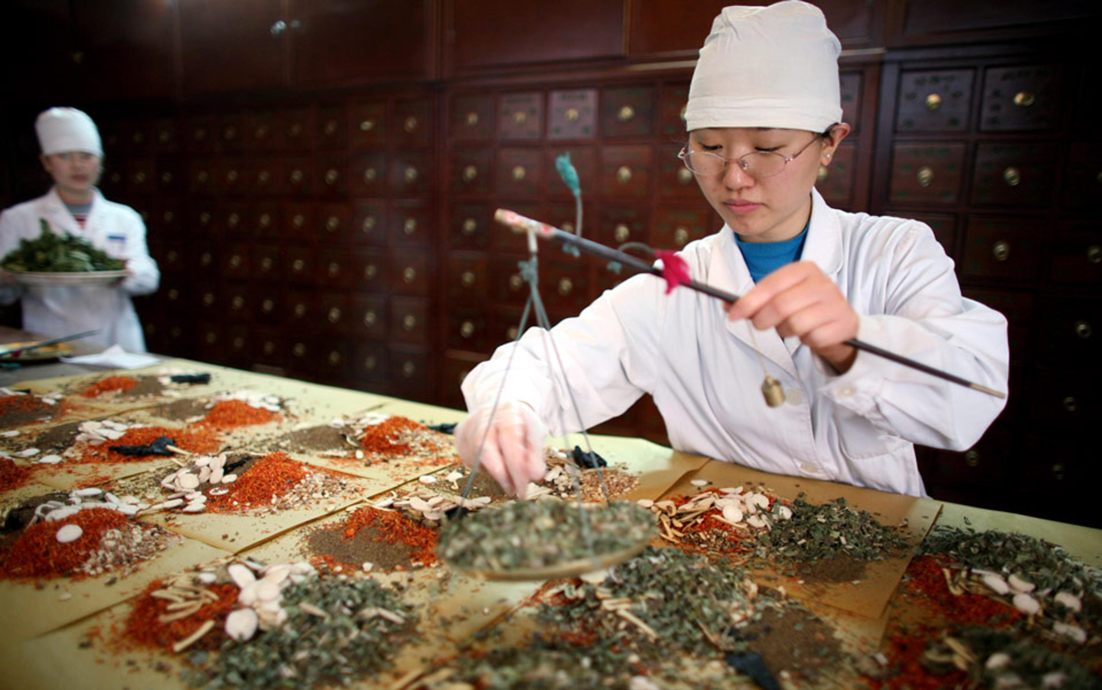 Two Chinese individuals in white coats and hats, preparing various herbs and traditional medicines on a table with small drawers behind.