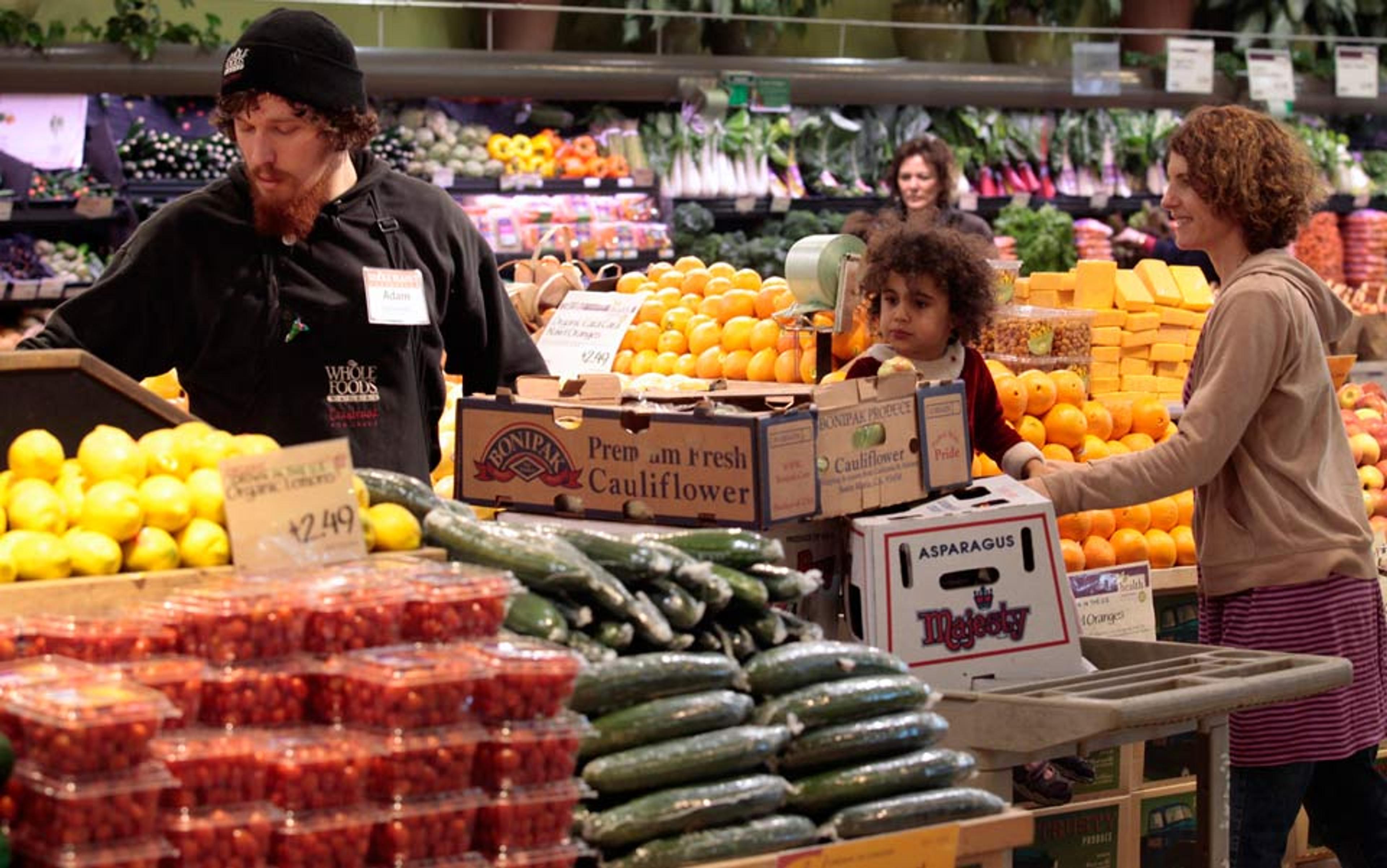 A market scene with a bearded man stocking produce near a smiling woman and child selecting items surrounded by fresh fruits and vegetables.