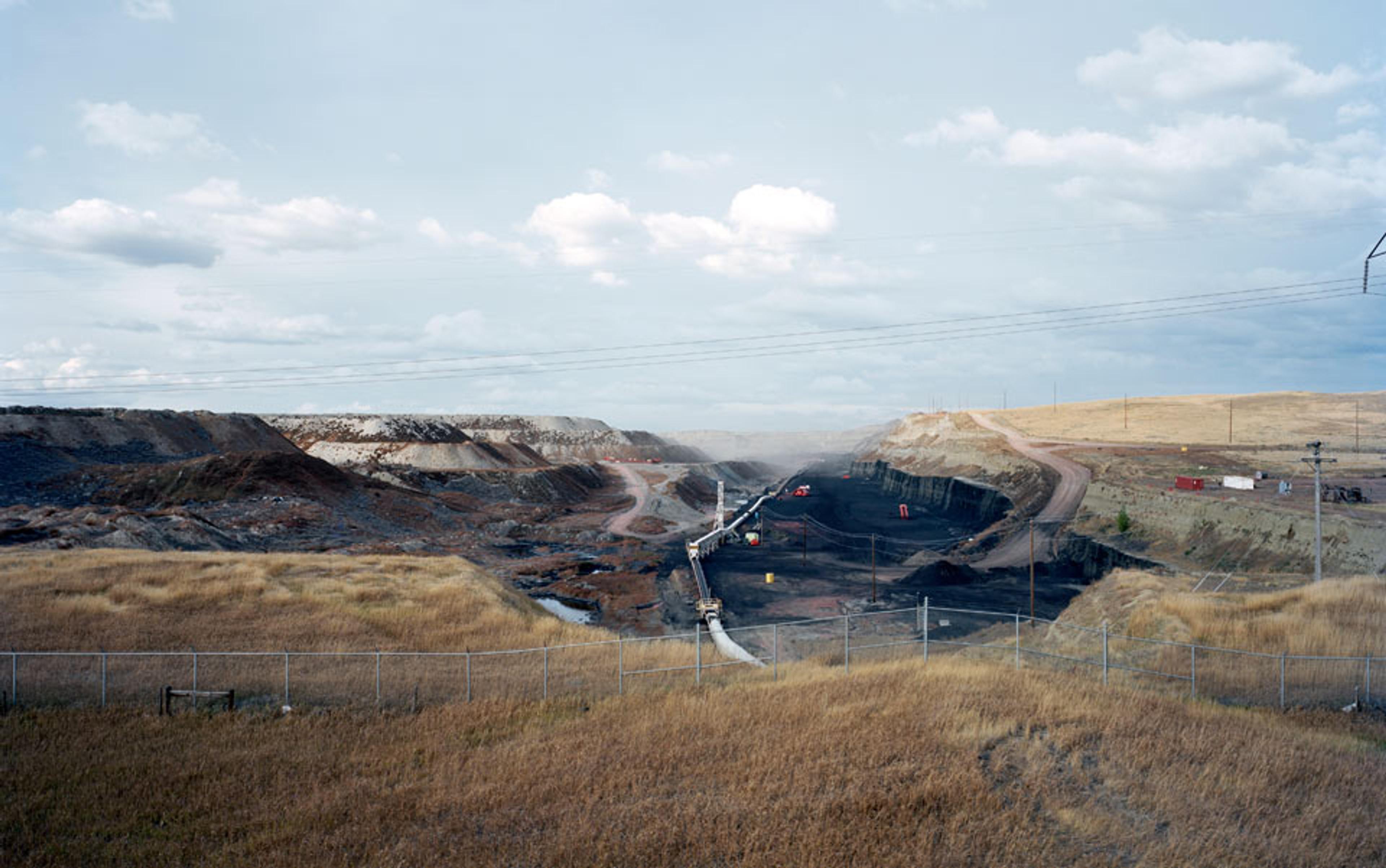 A large, open-pit coal mine with visible machinery and infrastructure, surrounded by dry grass and a fenced perimeter, under a cloudy sky.