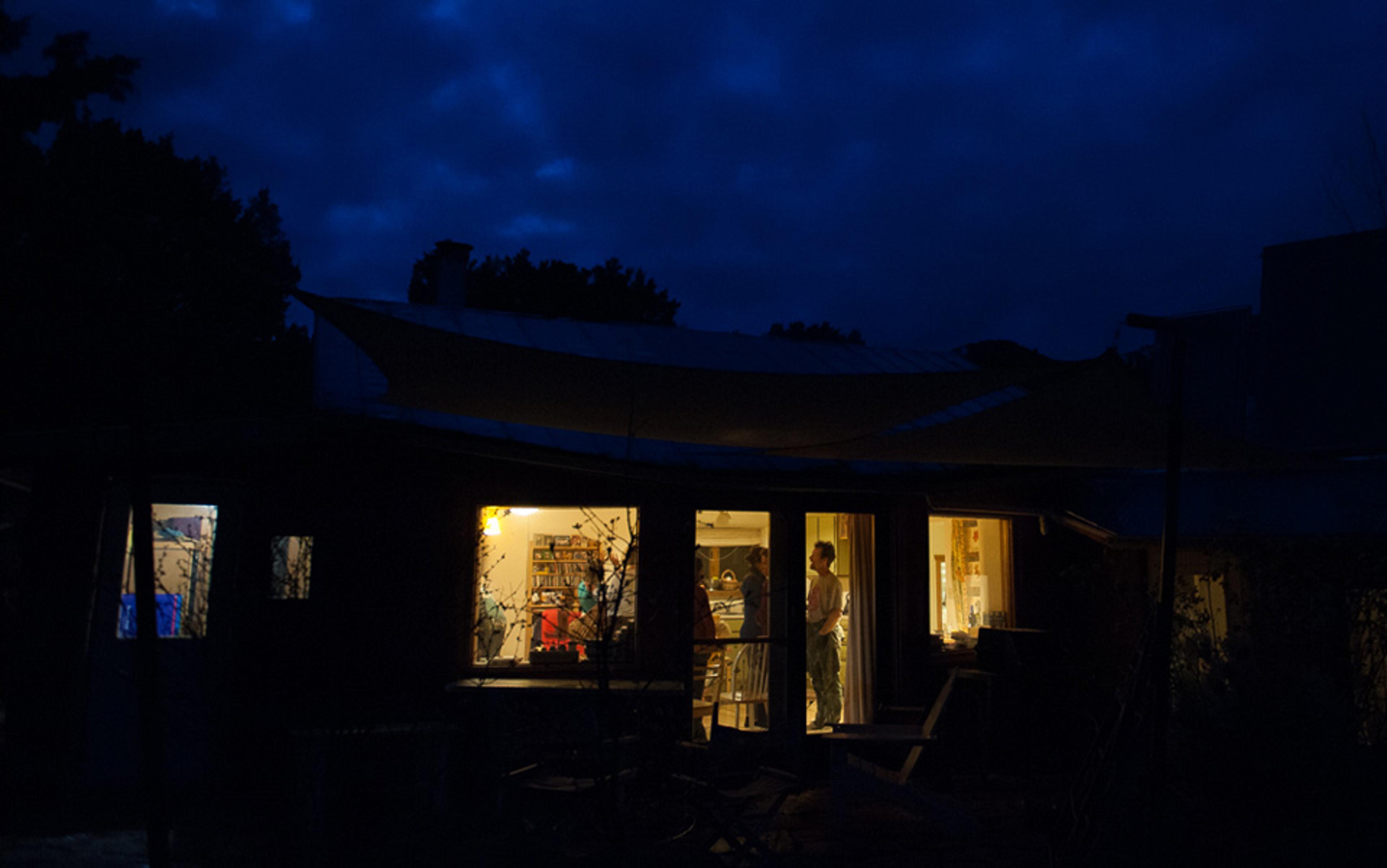 A house at night with lit interior showing people in the kitchen seen through large windows, surrounded by a dark exterior.